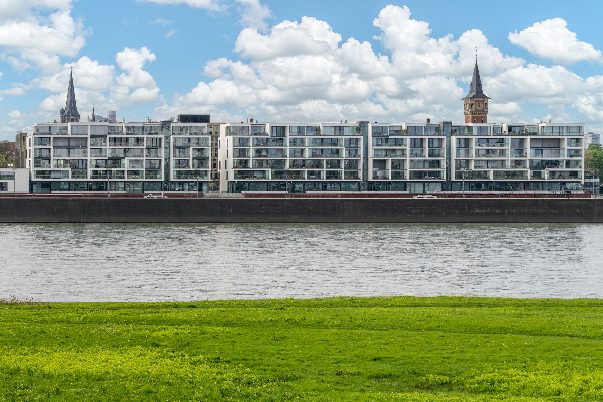 Modernes Wohngebäude mit vielen Balkonen am Flussufer, davor grüne Wiese und Wasser.