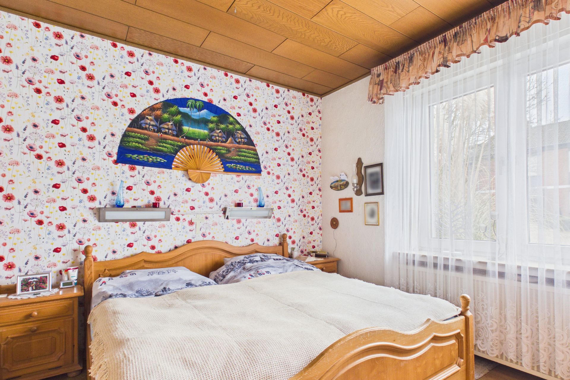 Helles Schlafzimmer mit Doppelbett, Blumentapete, Wandfächer und großem Fenster mit Vorhängen.