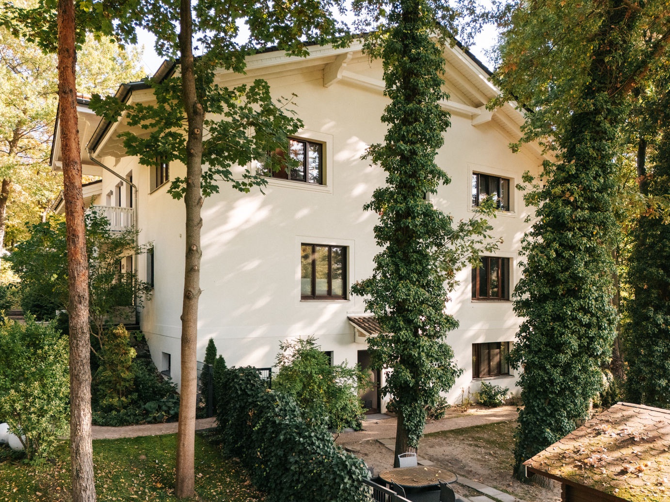 Weißes Wohnhaus mit vielen Fenstern, umgeben von hohen Bäumen und Garten.