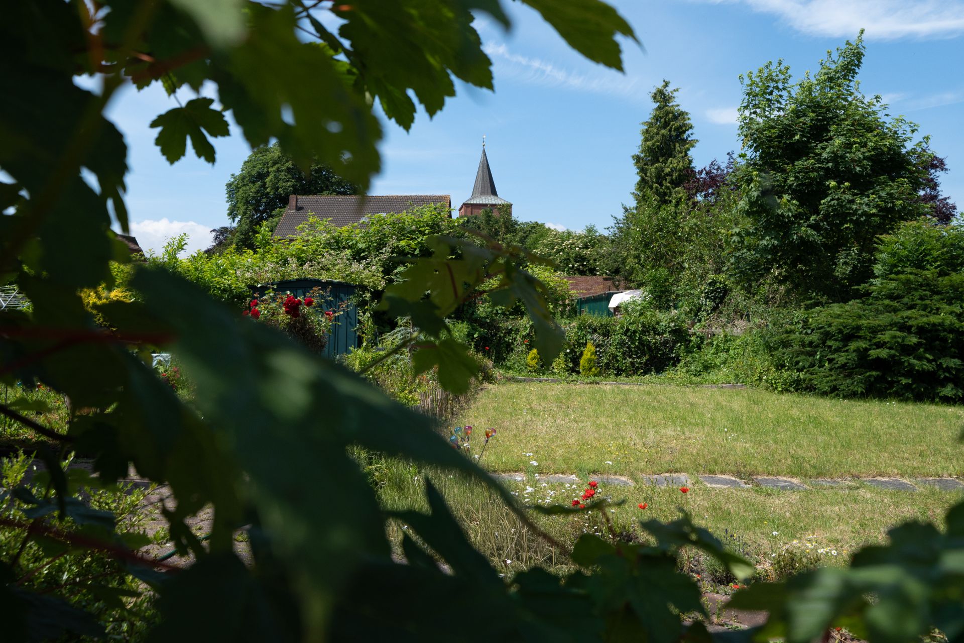 Garten mit Rasenfläche, Sträuchern und Blick auf einen Kirchturm im Hintergrund.
