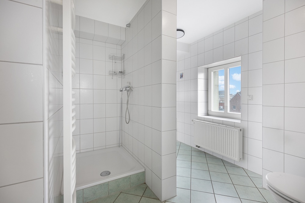 Helles Badezimmer mit Dusche, Heizkörper, Fenster und weißen Fliesen an Wand und Boden.