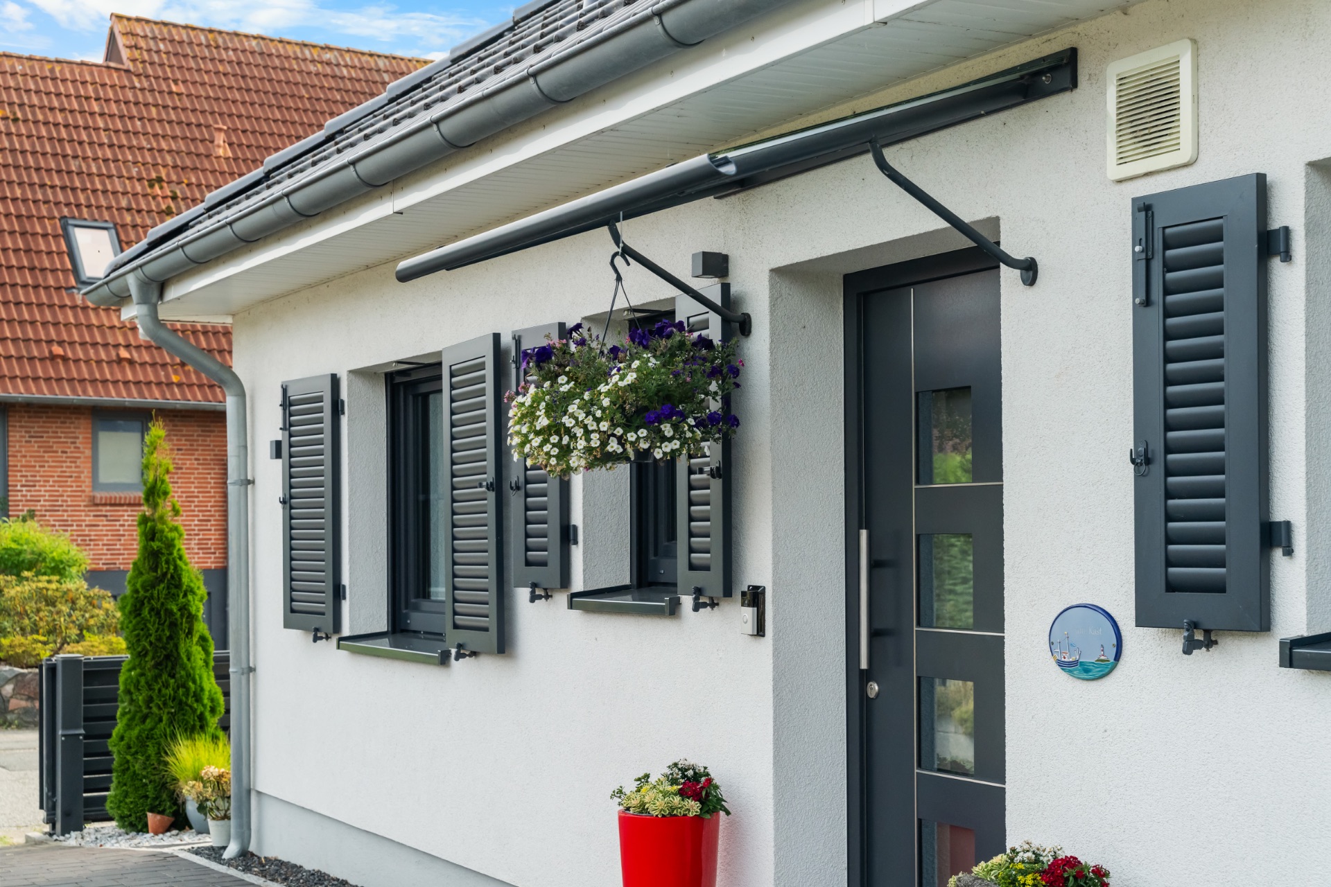 Weiße Hausfassade mit grauen Fensterläden, Hängkorb mit Blumen und rotem Blumentopf neben der Tür.