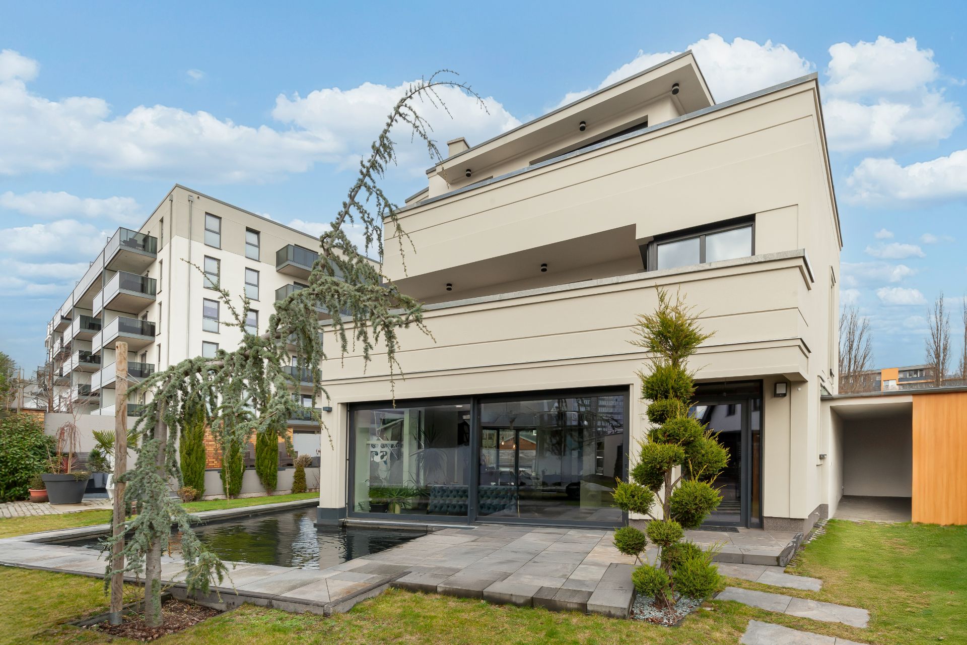 Moderne Wohnhausfassade mit großen Fenstern, Terrasse, Teich und gepflegtem Garten.