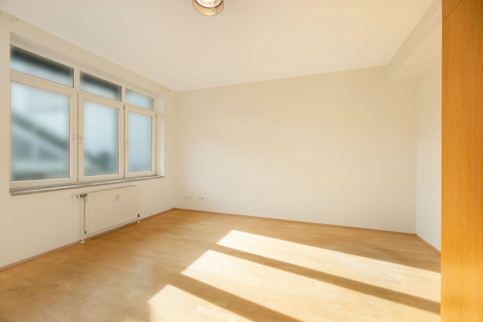 Heller, leerer Raum mit großem Fenster, Parkettboden und weißen Wänden.