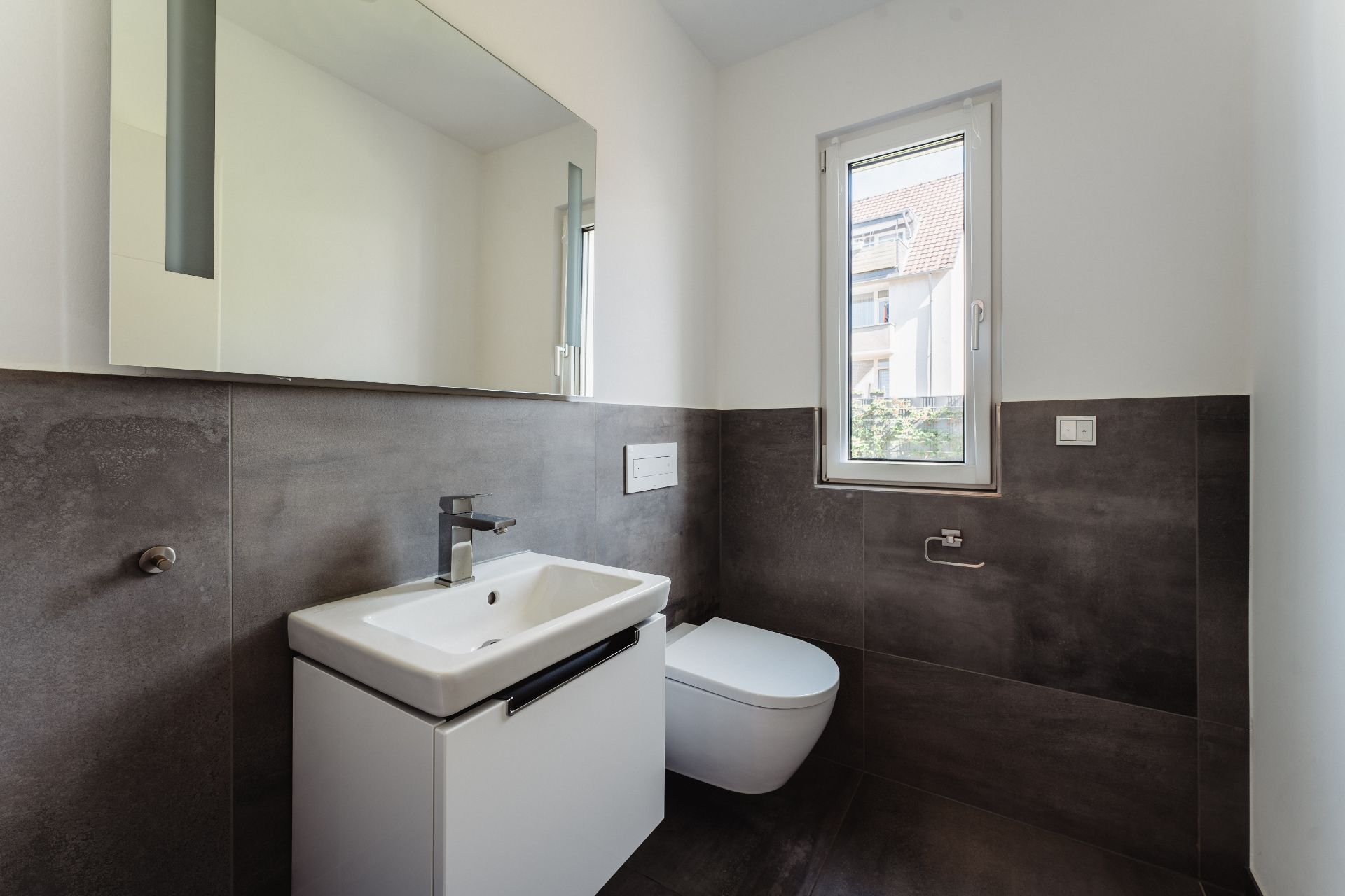 Modernes Badezimmer mit Waschbecken, Spiegel, WC und Fenster, dunkle Fliesen an der Wand.