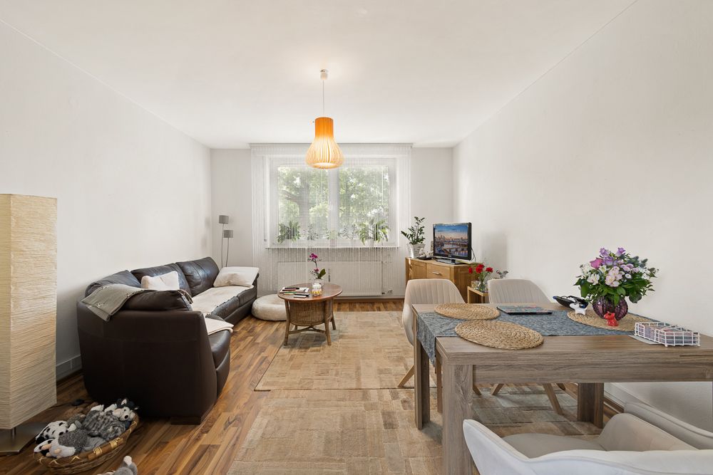 Helles Wohnzimmer mit Sofa, Esstisch, Teppichboden und großen Fenstern.