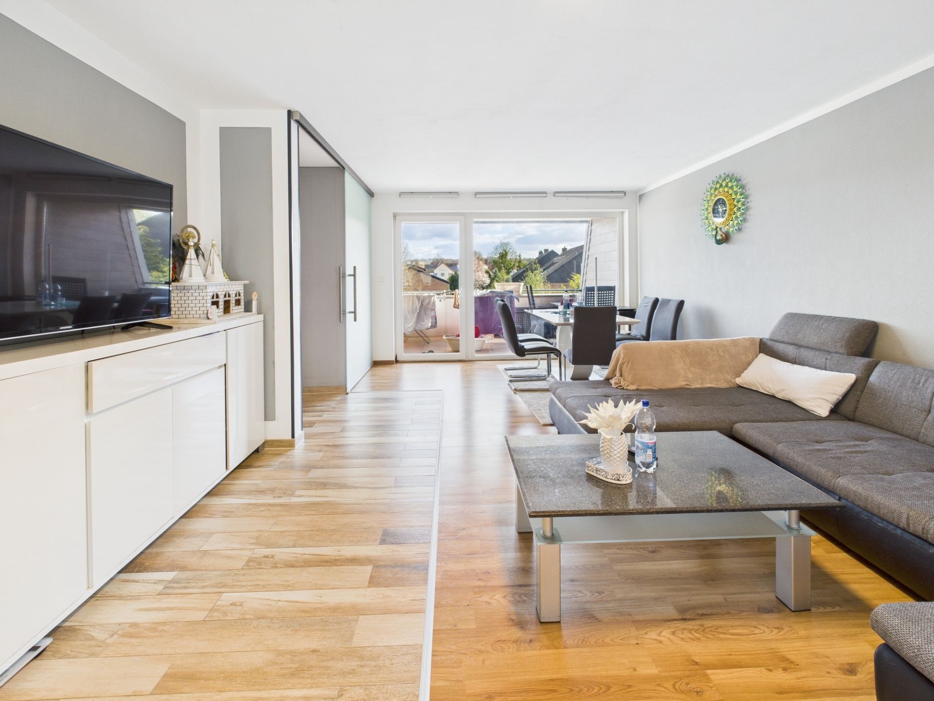 Helles Wohnzimmer mit grauem Sofa, Couchtisch, großem Fernseher und Essbereich, Glastür zum Balkon.