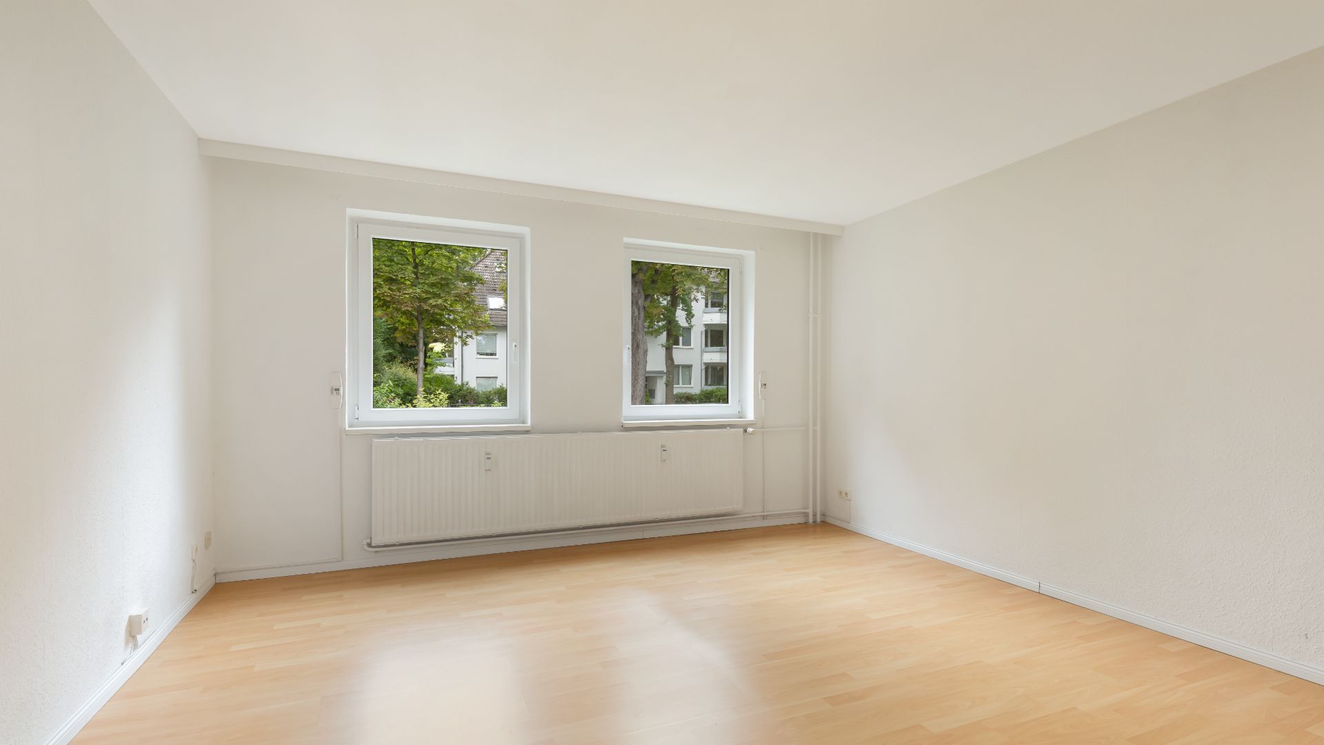 Helles, leeres Zimmer mit zwei Fenstern, weiß gestrichenen Wänden und hellem Holzfußboden.