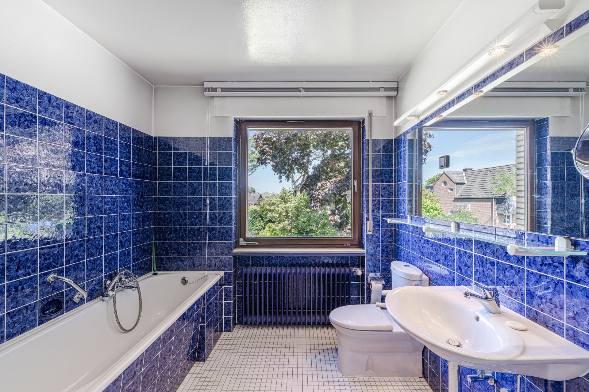Badezimmer mit blauen Fliesen, Badewanne, WC, Waschbecken und Fenster mit Ausblick.