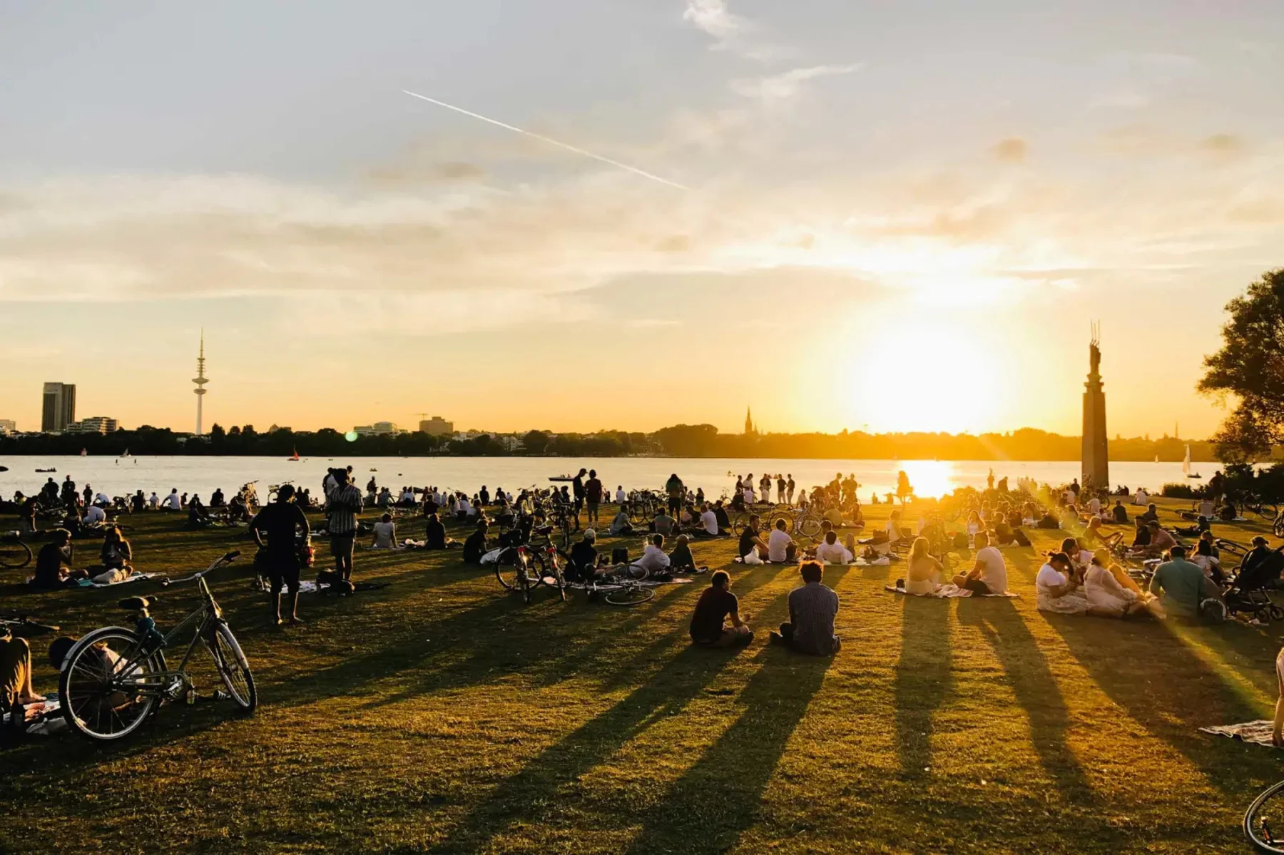 Viele Menschen sitzen auf einer Wiese am Wasser bei Sonnenuntergang.