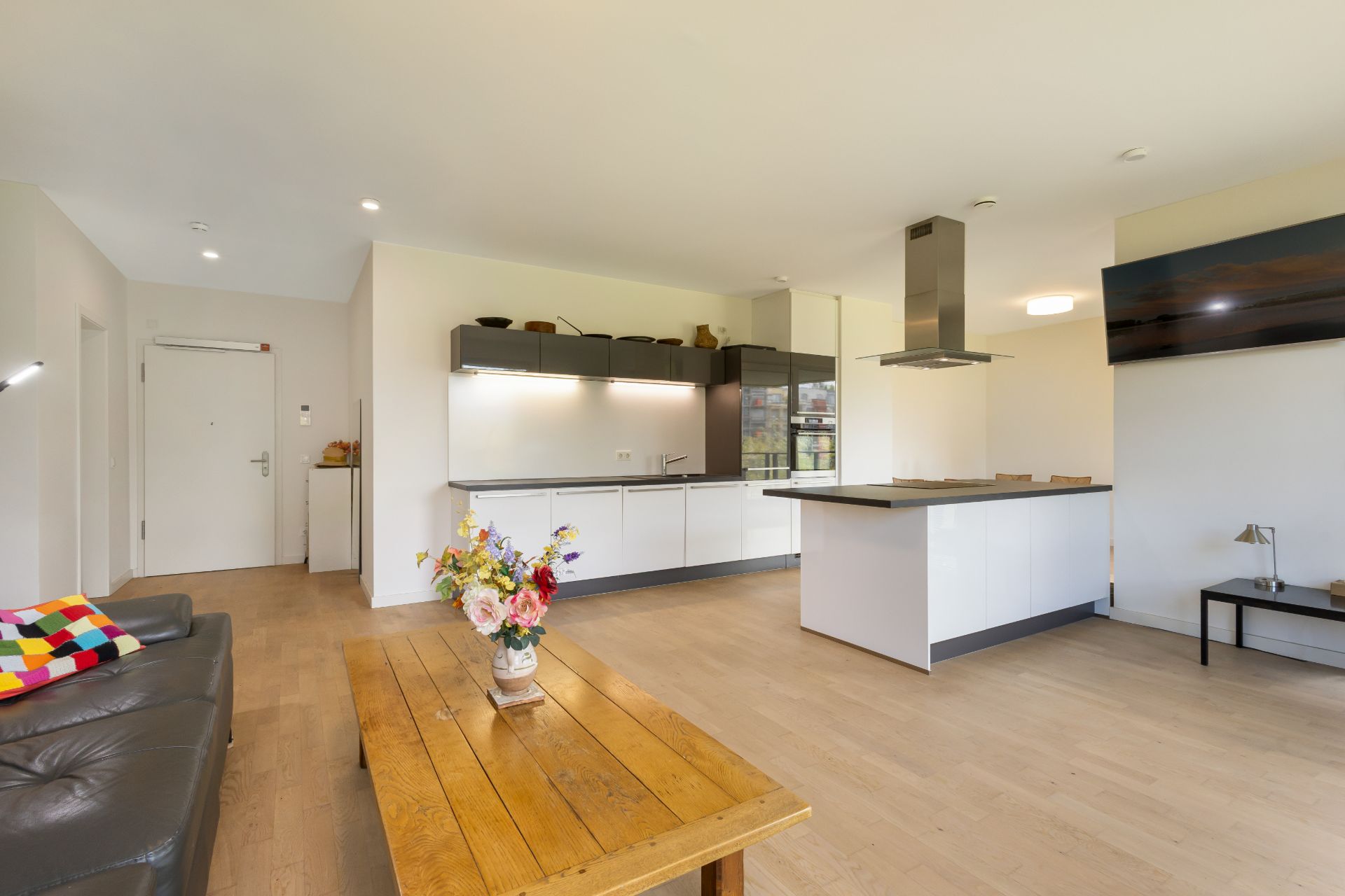 Helles Wohnzimmer mit offener Küche, Holztisch, Sofa und Blumenstrauß auf dem Tisch.