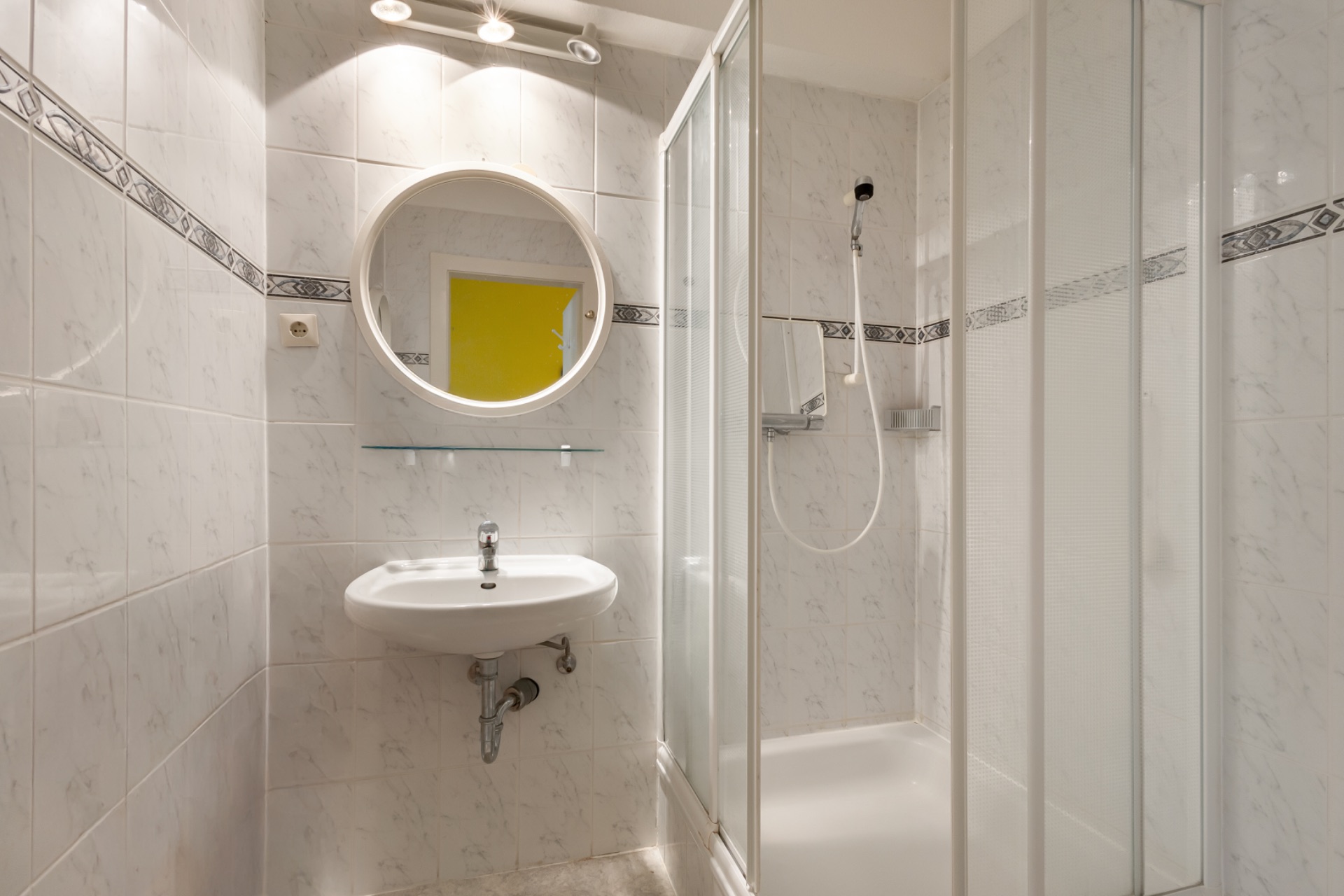 Kleines Badezimmer mit Dusche, rundem Spiegel und Waschbecken, gefliest in Weiß.