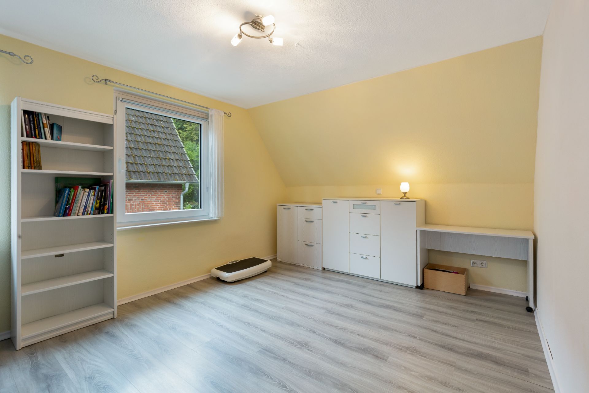 Helles Zimmer mit Bücherregal, Schränken, Schreibtisch und Fenster, Laminatboden.