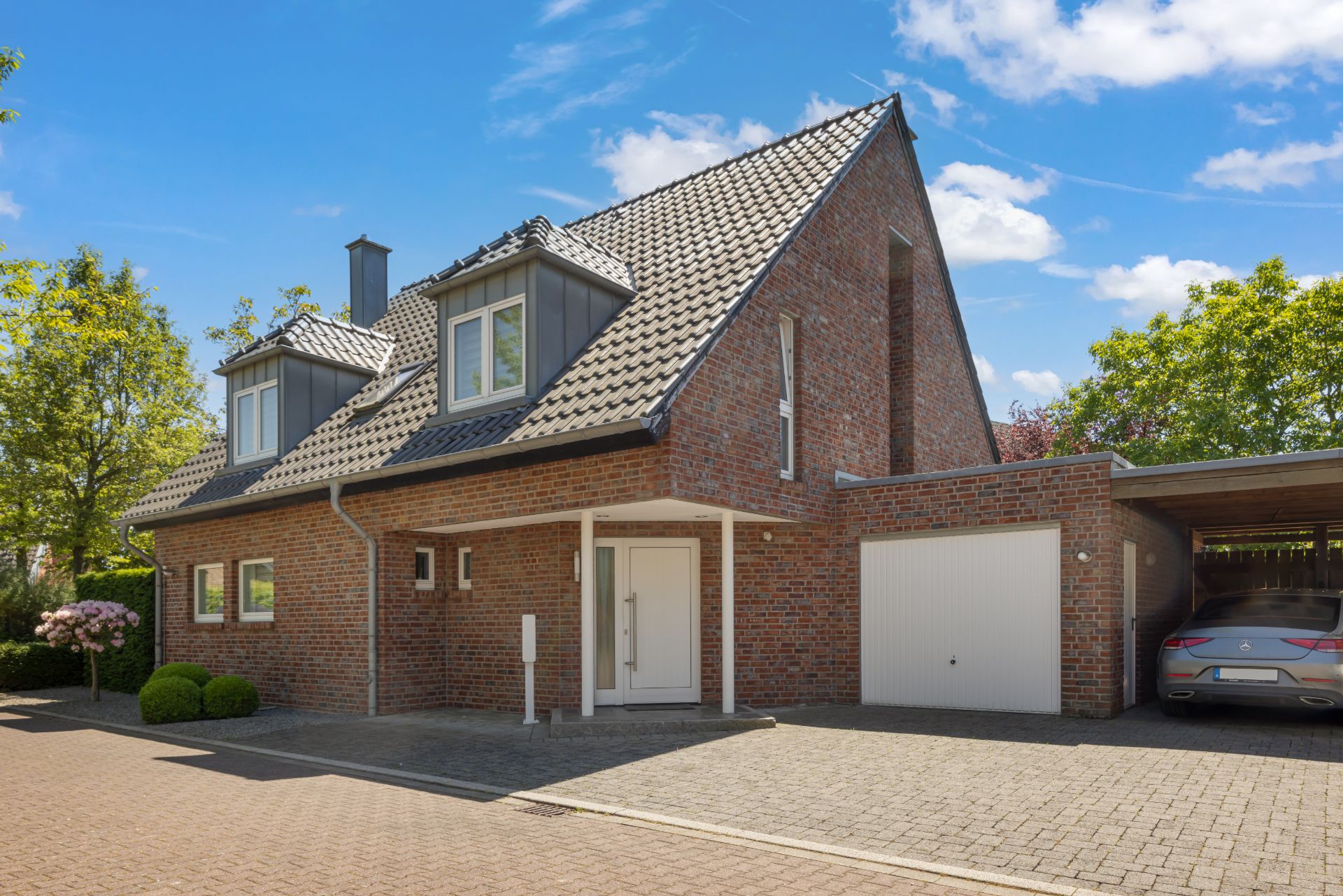 Backsteinhaus mit Garage, Carport und auffahrendem Auto, umgeben von Bäumen.