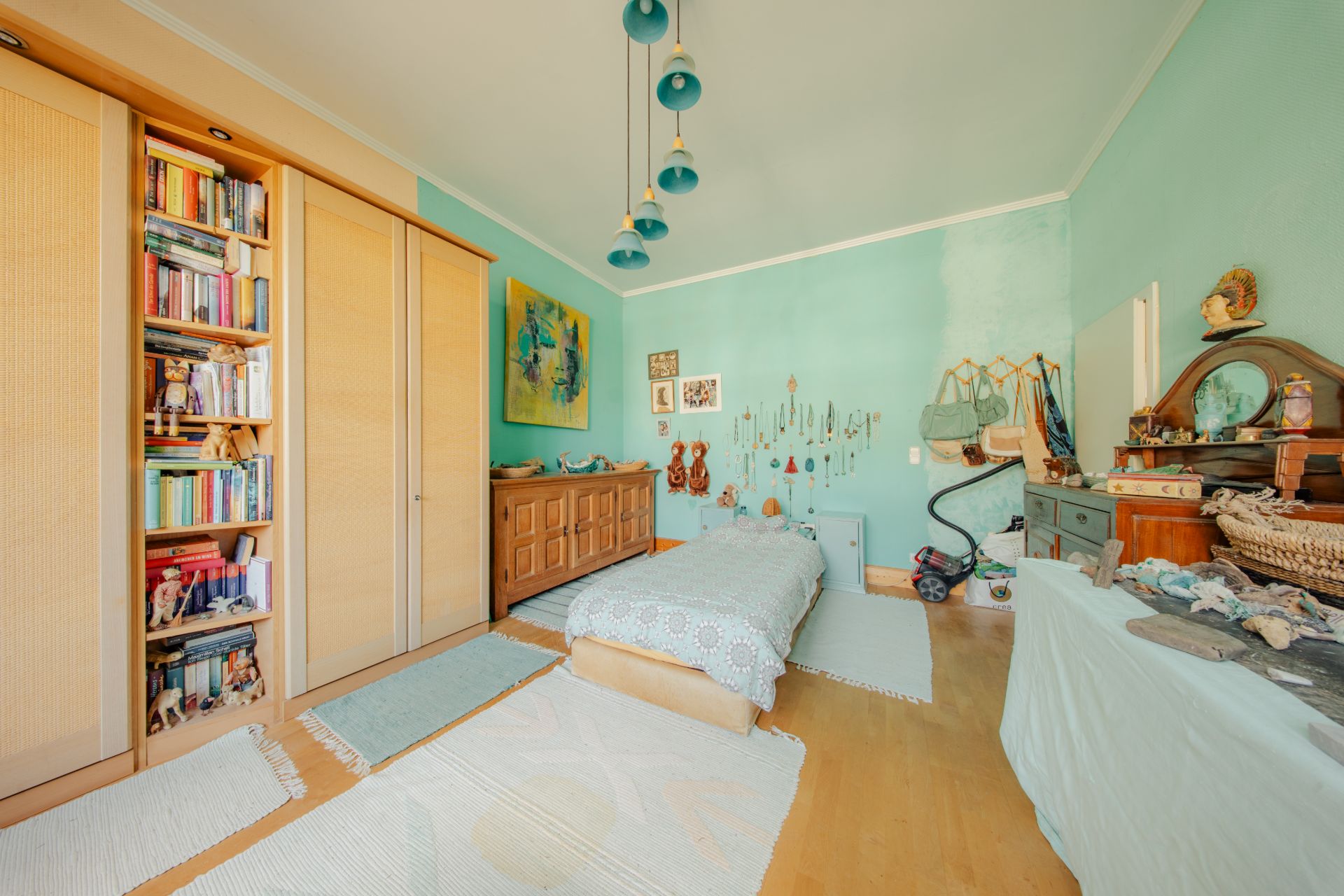Helles Schlafzimmer mit Einzelbett, Holzschränken, Bücherregal und türkisfarbenen Wänden.