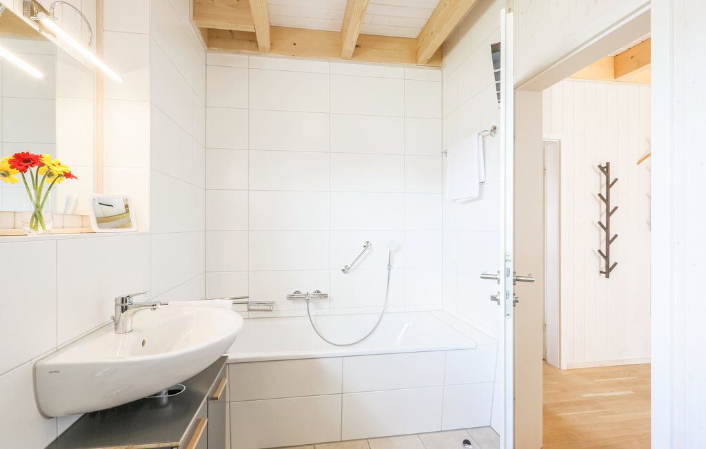 Helles Bad mit weiß gefliester Badewanne, Waschbecken und Holzdecke, Vase mit Blumen auf dem Spiegelregal.