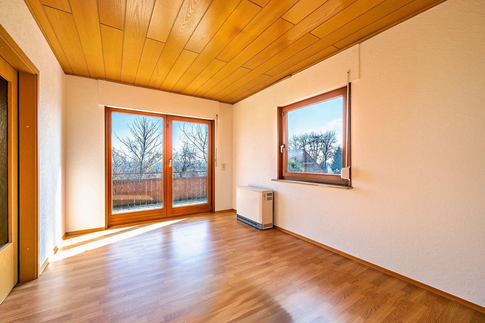 Heller Raum mit Holzdecke, Parkettboden, Fenster und Glastür zum Balkon.