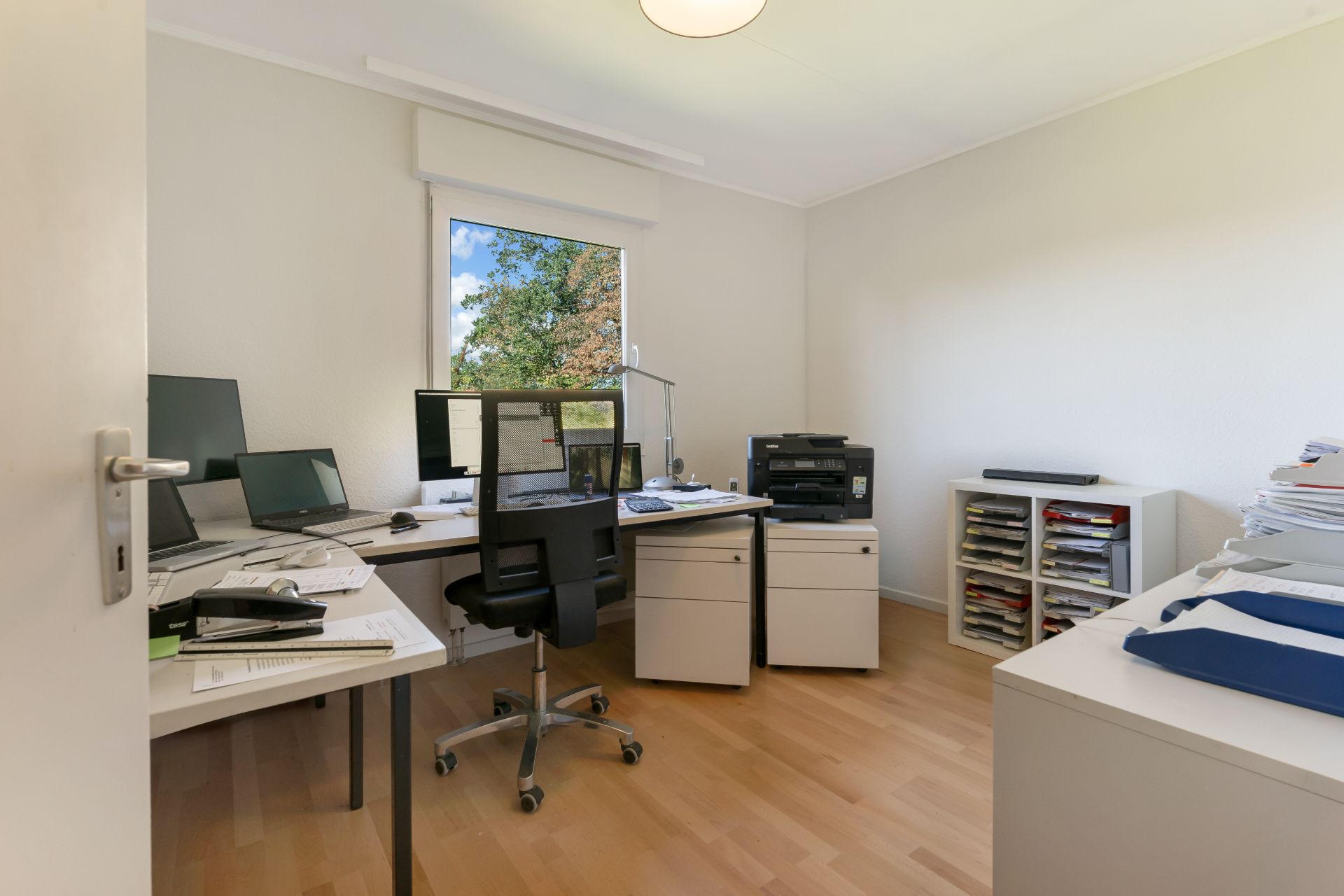 Helles Arbeitszimmer mit Schreibtischen, mehreren Monitoren, Stuhl und Regalen vor einem Fenster.
