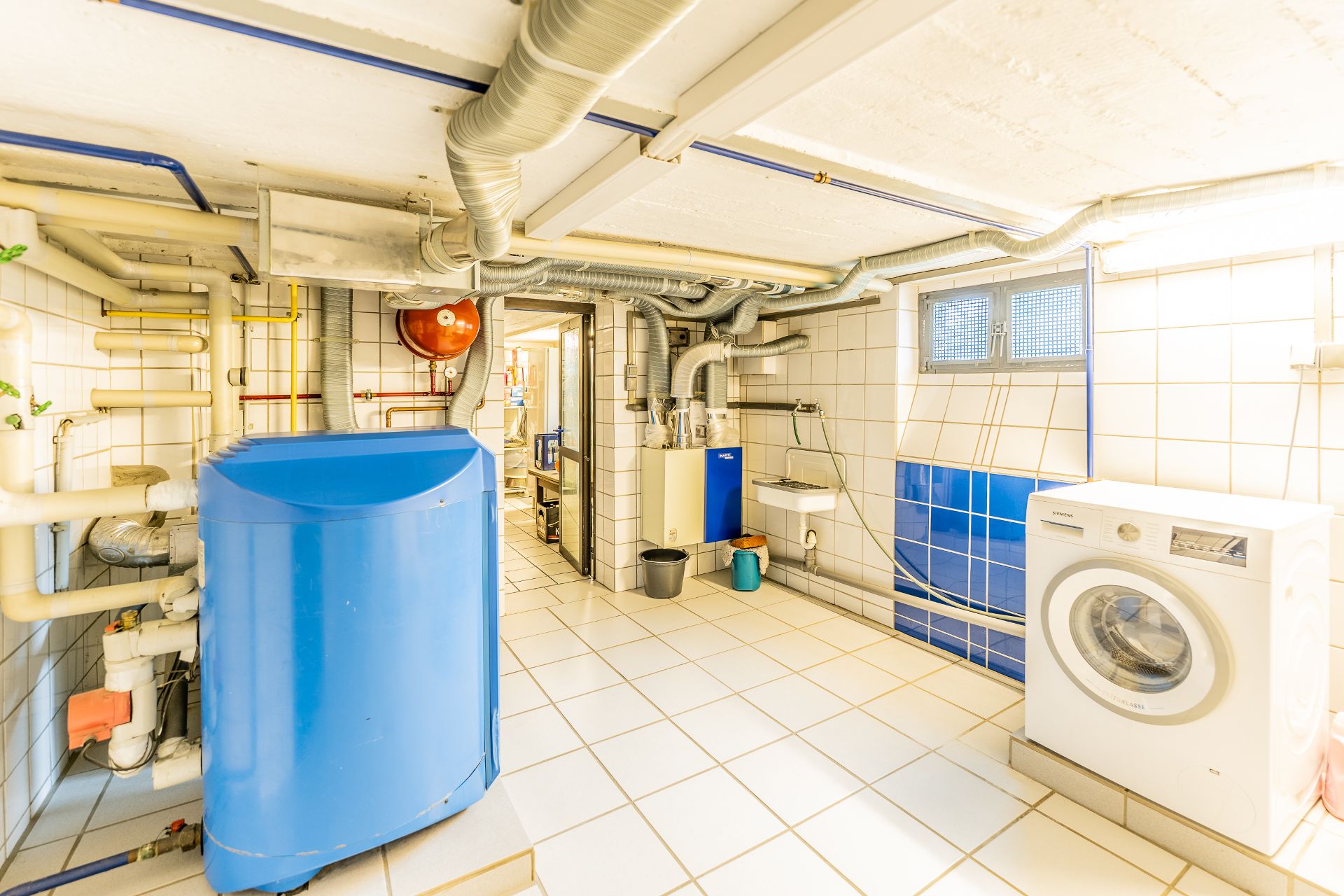 Heller Hauswirtschaftsraum mit Waschmaschine, Heizungsanlage, Rohren und kleinem Fenster.