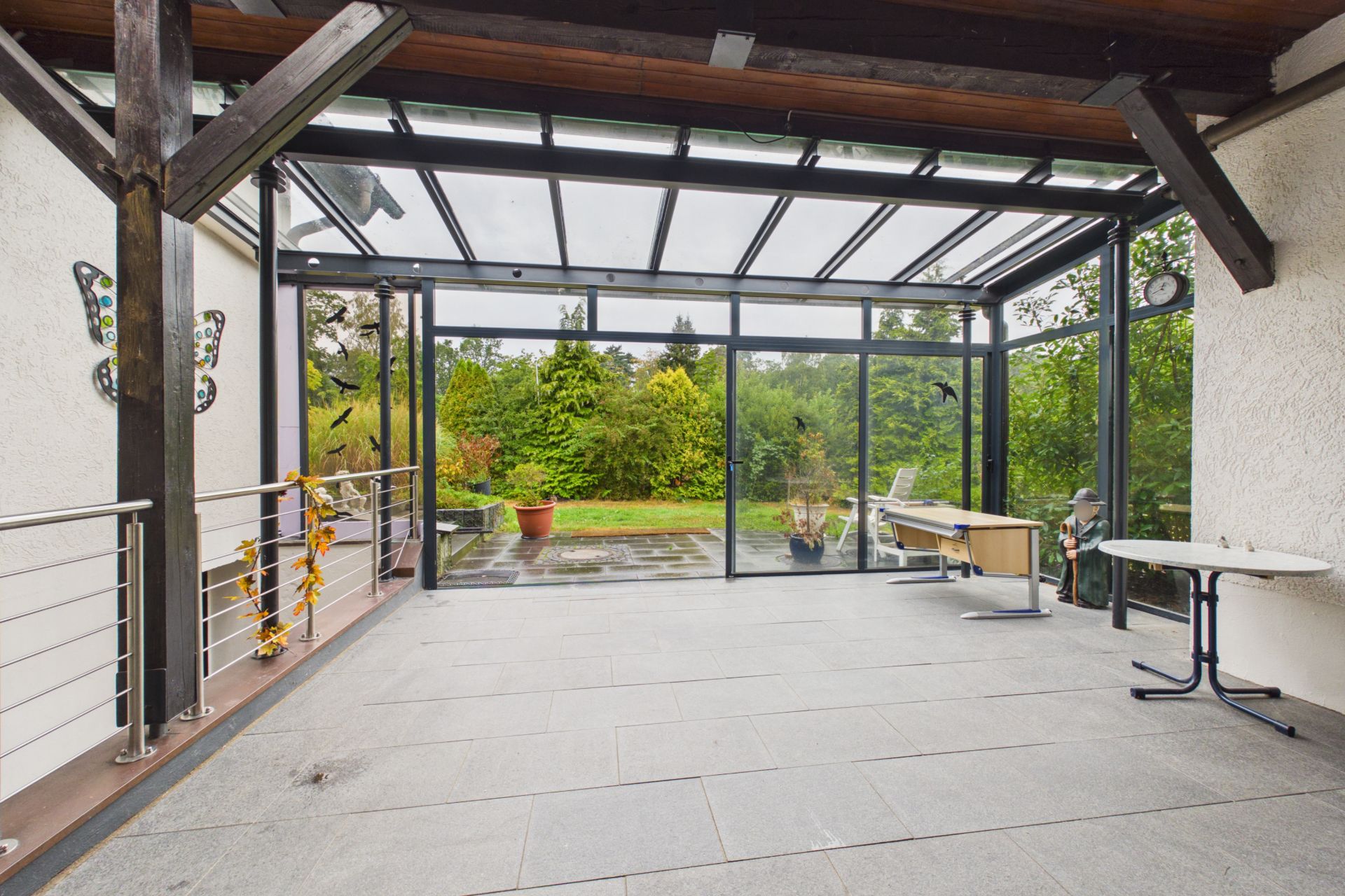 Überdachte Terrasse mit Glasdach, Blick auf Garten, runder Tisch und Schreibtisch.