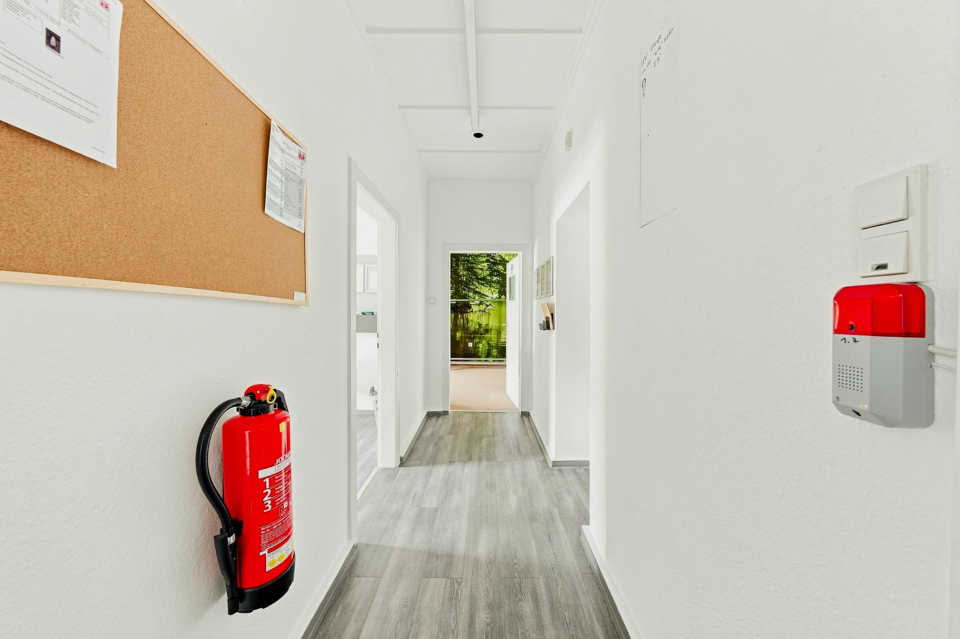 Heller Flur mit grauem Boden, Feuerlöscher und Pinnwand an der Wand.