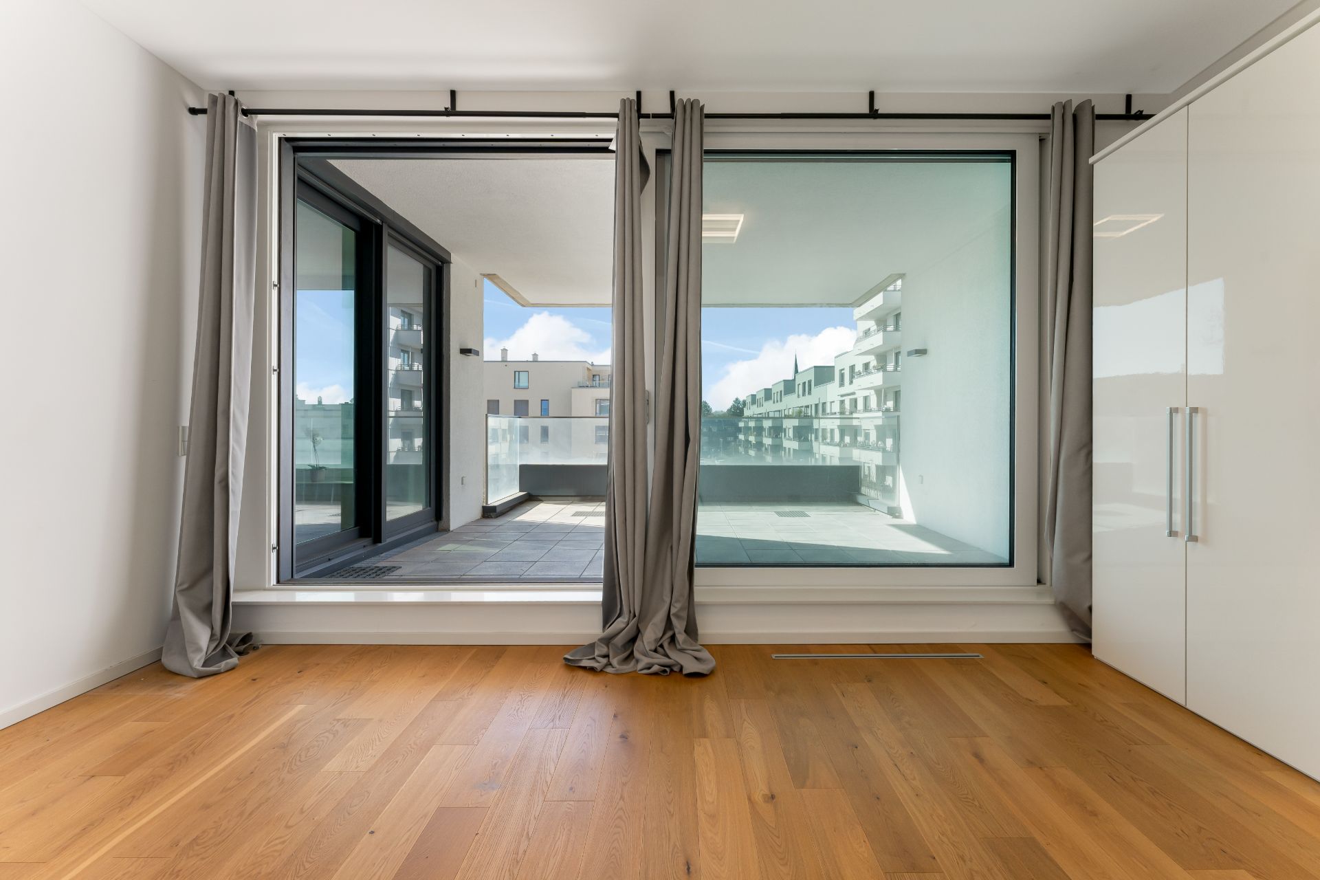 Helles Zimmer mit Holzboden, bodentiefen Fenstern, grauen Vorhängen und Zugang zu einem Balkon.