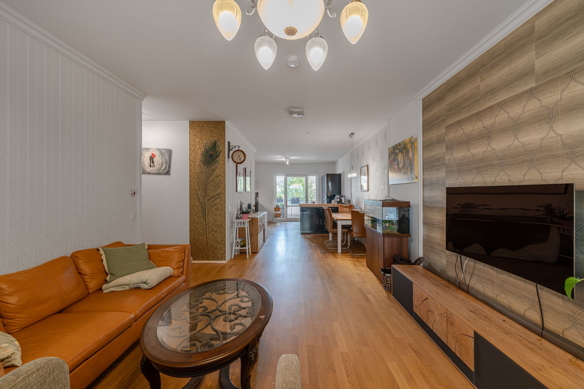 Modernes Wohnzimmer mit braunem Sofa, Glastisch, Fernseher und Essbereich im Hintergrund.