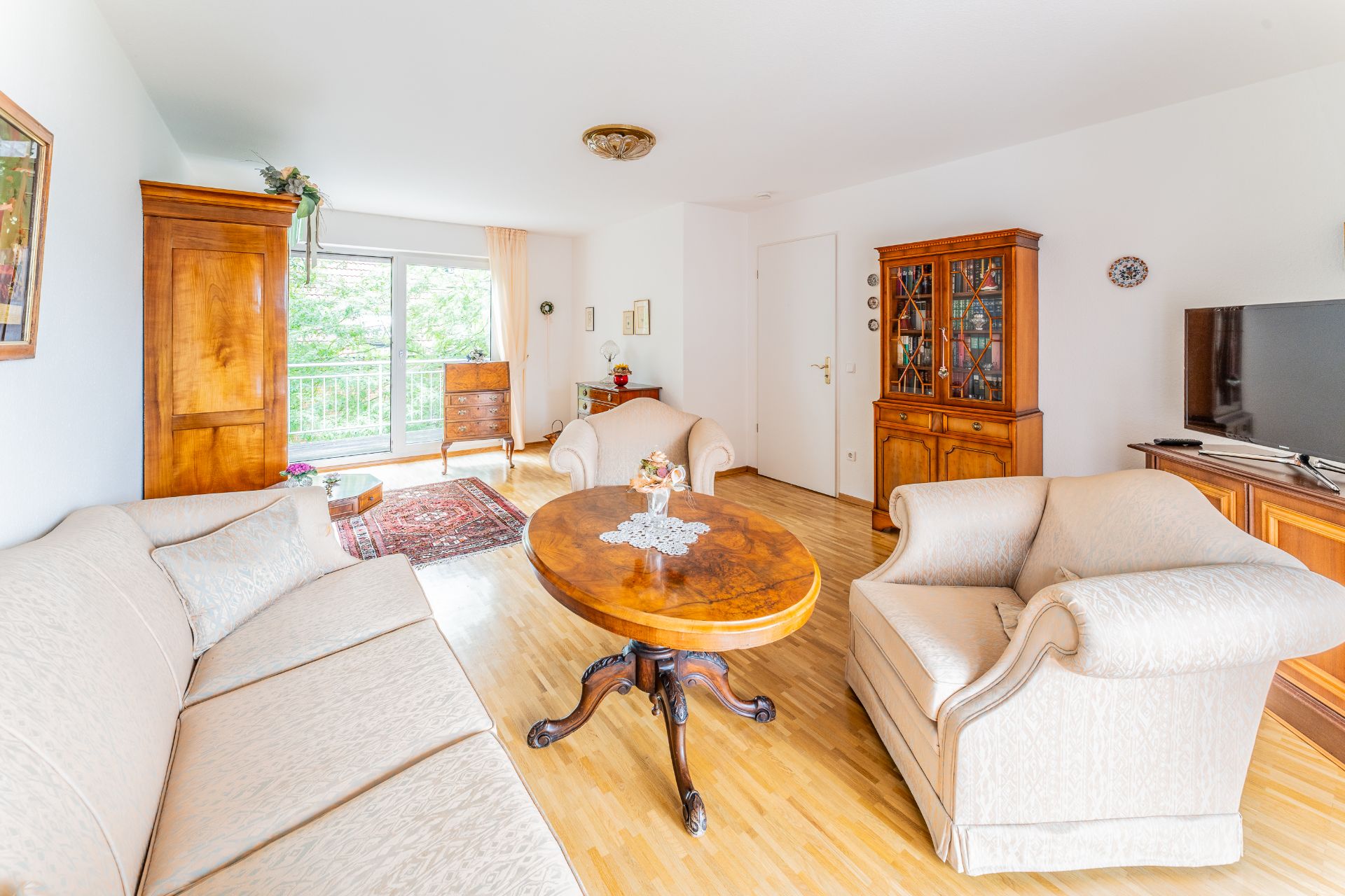 Helles Wohnzimmer mit cremefarbenen Polstermöbeln, Holztisch, Vitrine und Balkonblick.