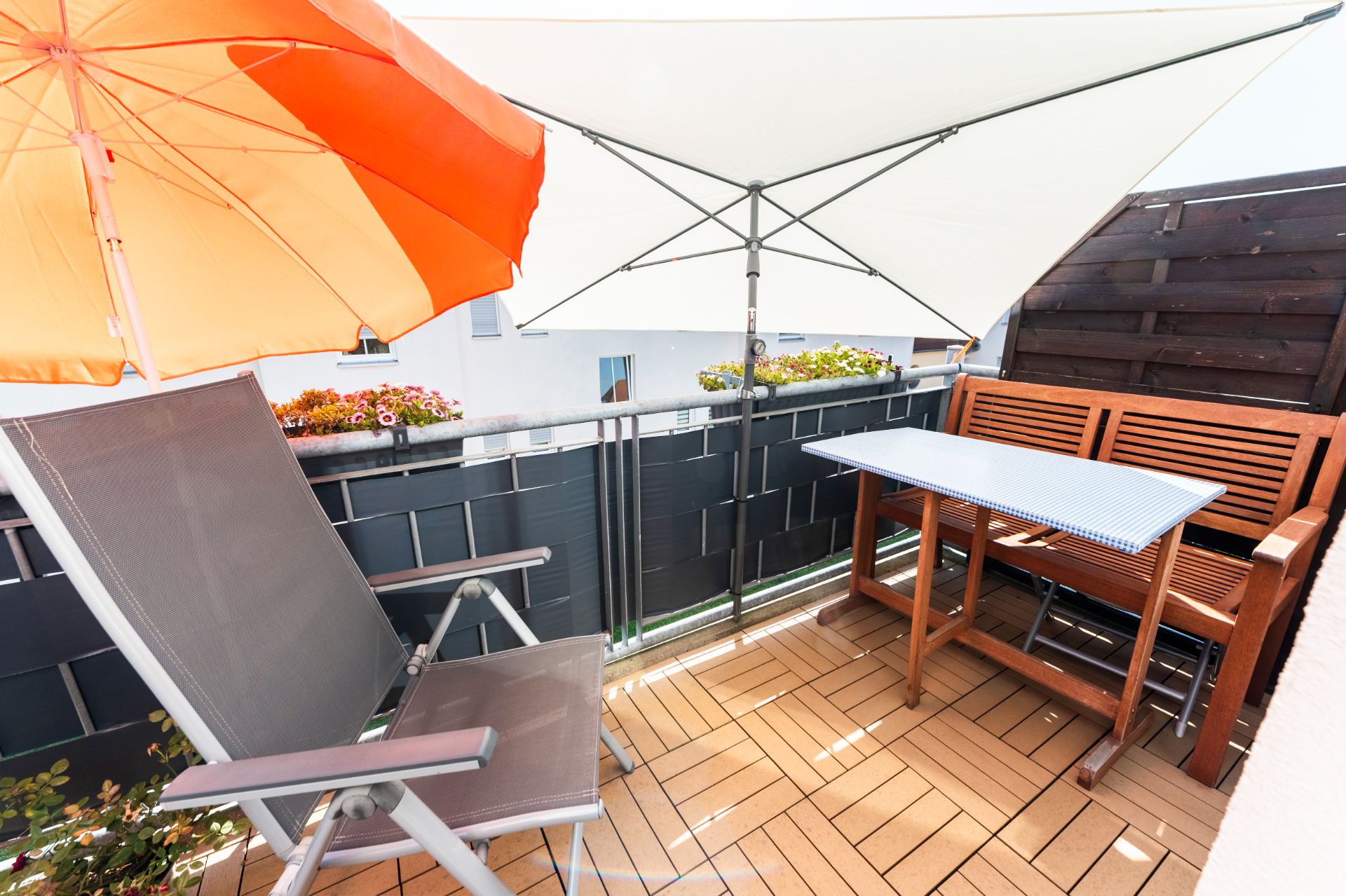Balkon mit Liegestuhl, Holztisch, Bank und Sonnenschirmen, Blumenkästen am Geländer.