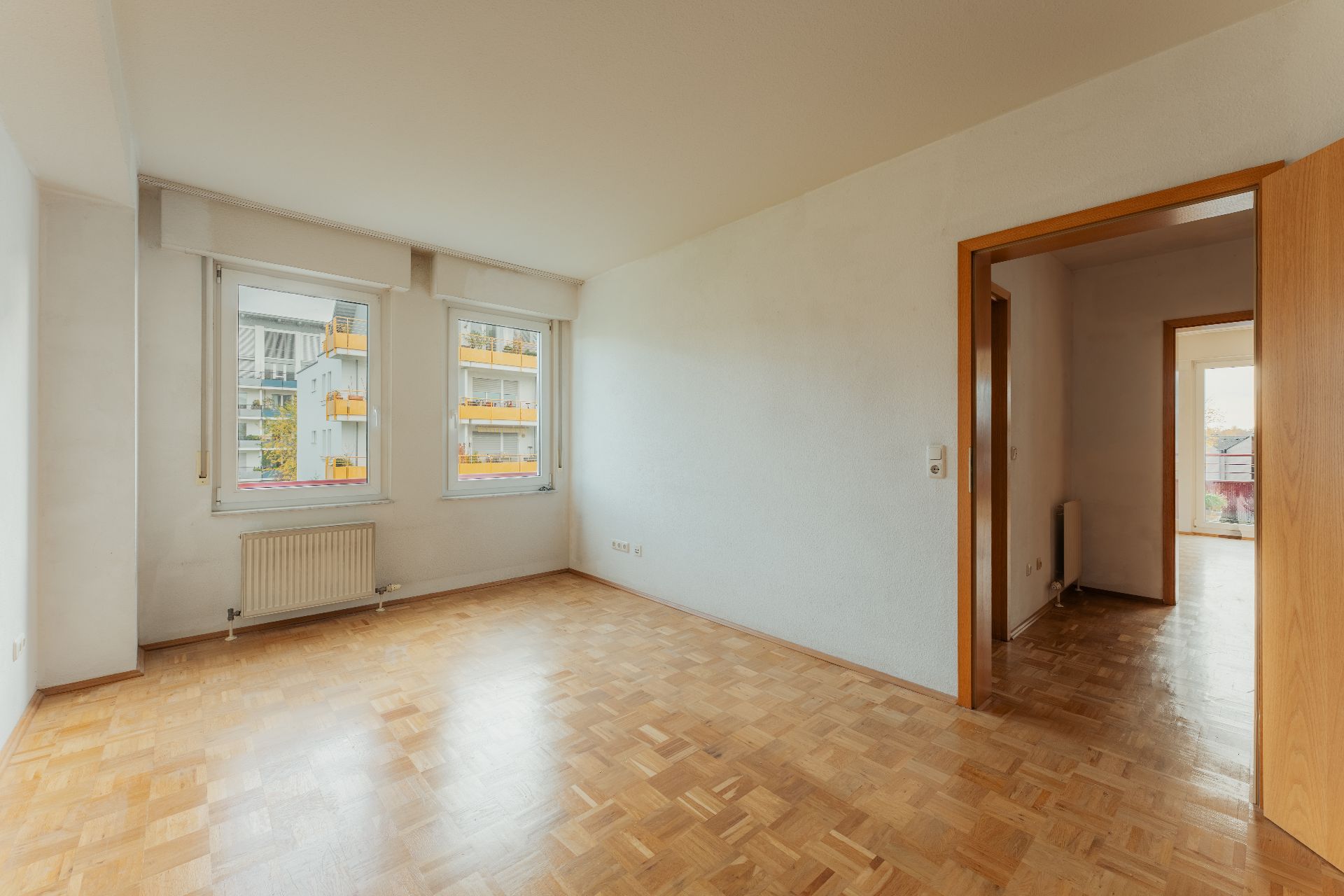 Helles Zimmer mit Parkettboden, zwei Fenstern und offenem Durchgang zu einem weiteren Raum.