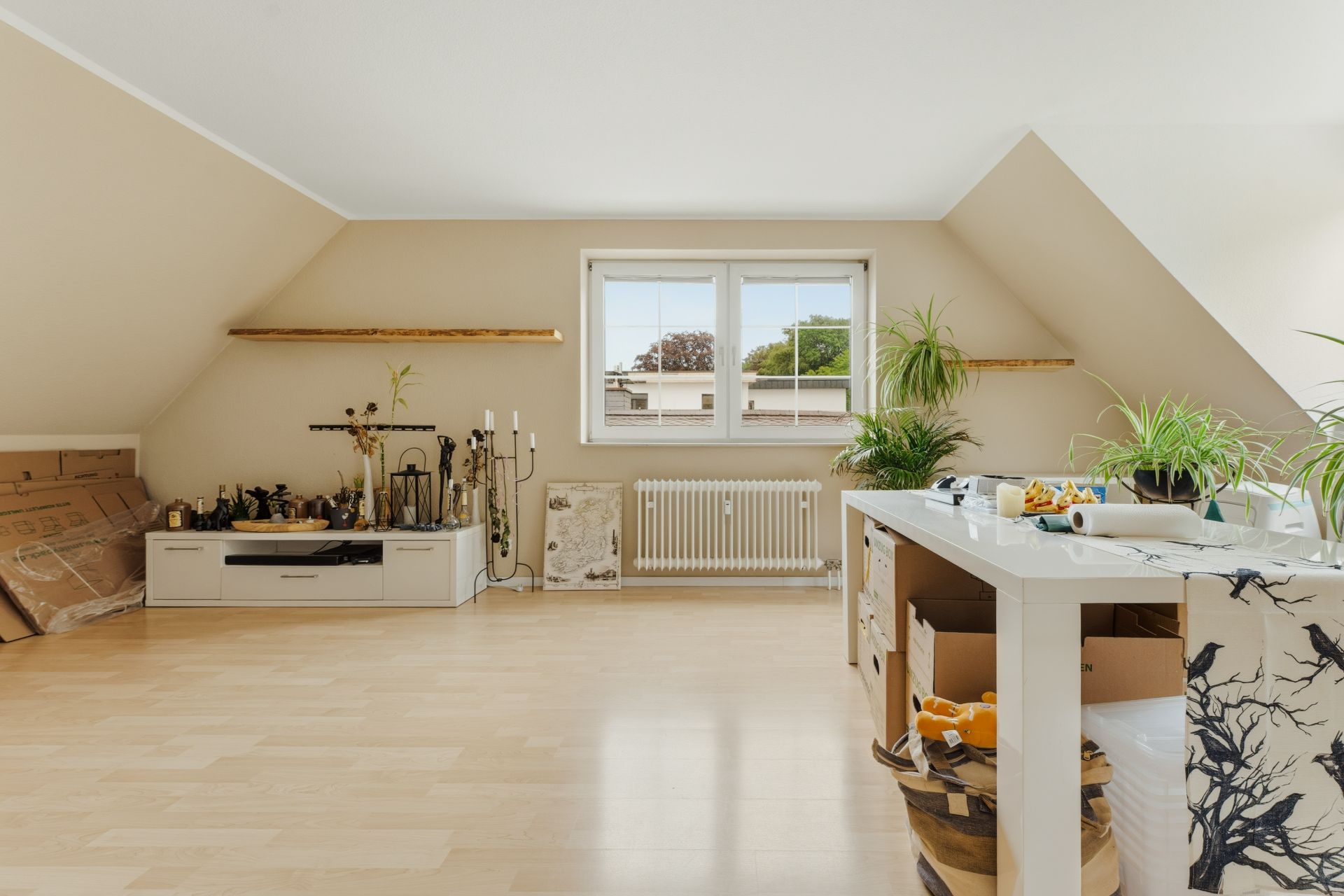 Helles Zimmer mit Dachschrägen, Fenster, Pflanzen und Tisch. Holzfußboden und offene Ablagen sind sichtbar.