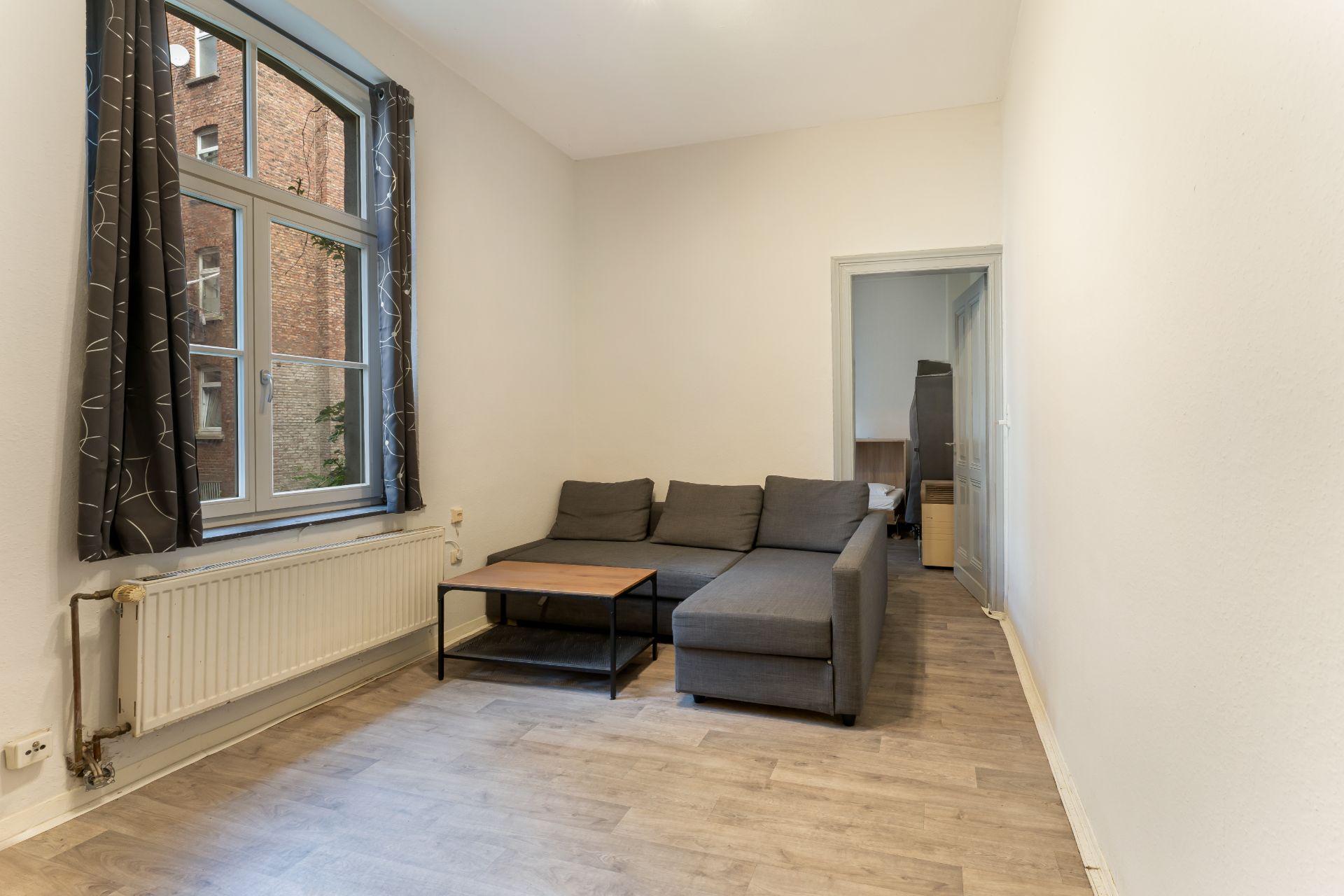 Wohnzimmer mit grauem Sofa, Couchtisch, großem Fenster und hellem Boden.
