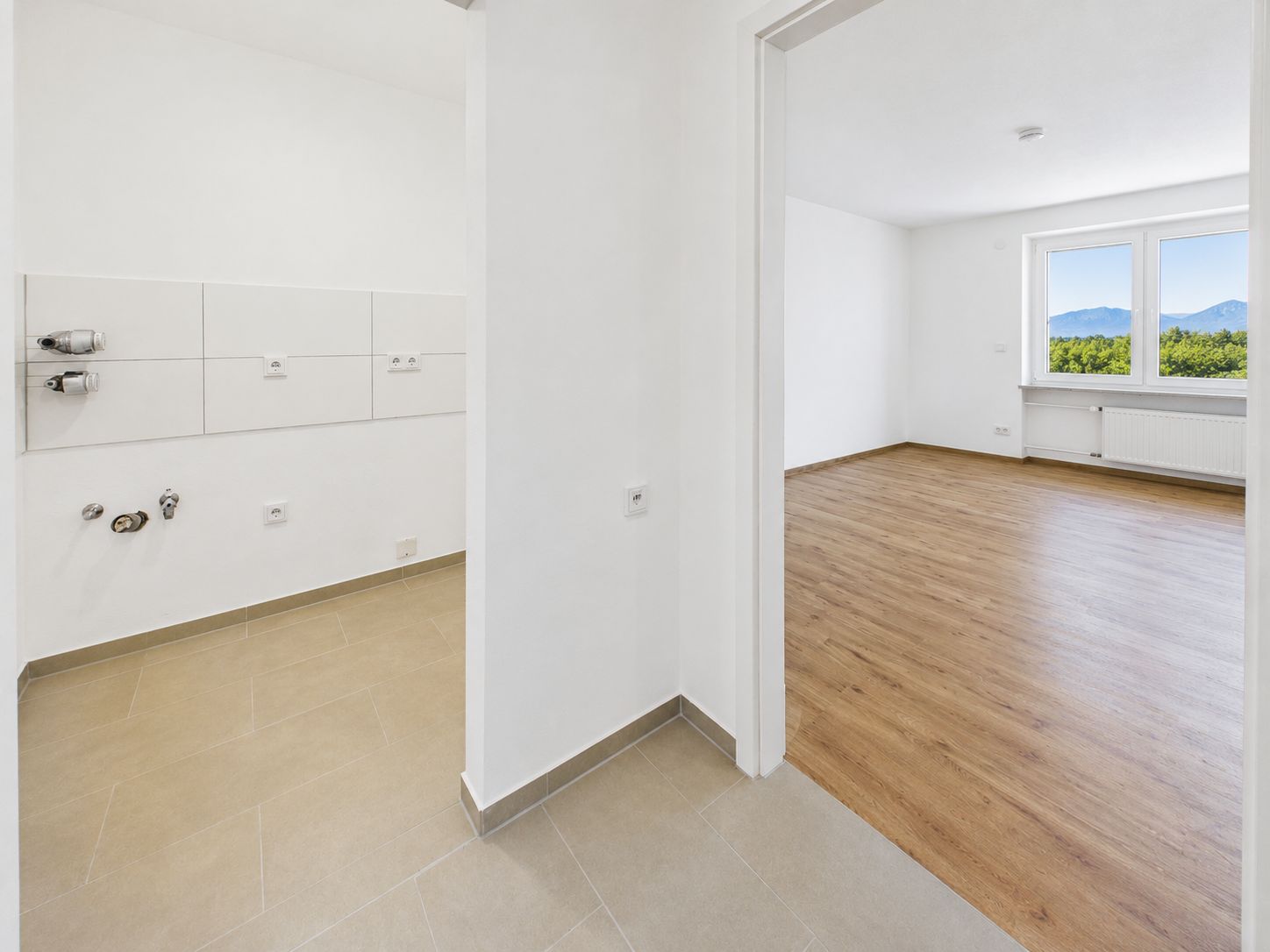 Heller Raum mit Holzfußboden und Fenster, daneben gefliester Raum mit Anschlüssen.