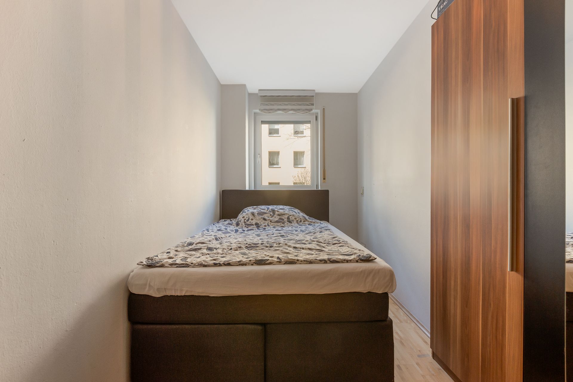 Schmaler Raum mit einem Bett, Fenster am Ende und einem Kleiderschrank rechts.