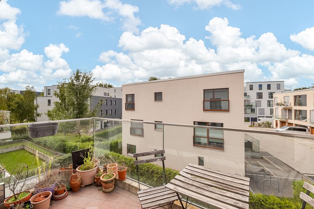 Balkon mit Holztisch, Stuhl, Blumentöpfen und Glasgeländer, Blick auf moderne Wohnhäuser.