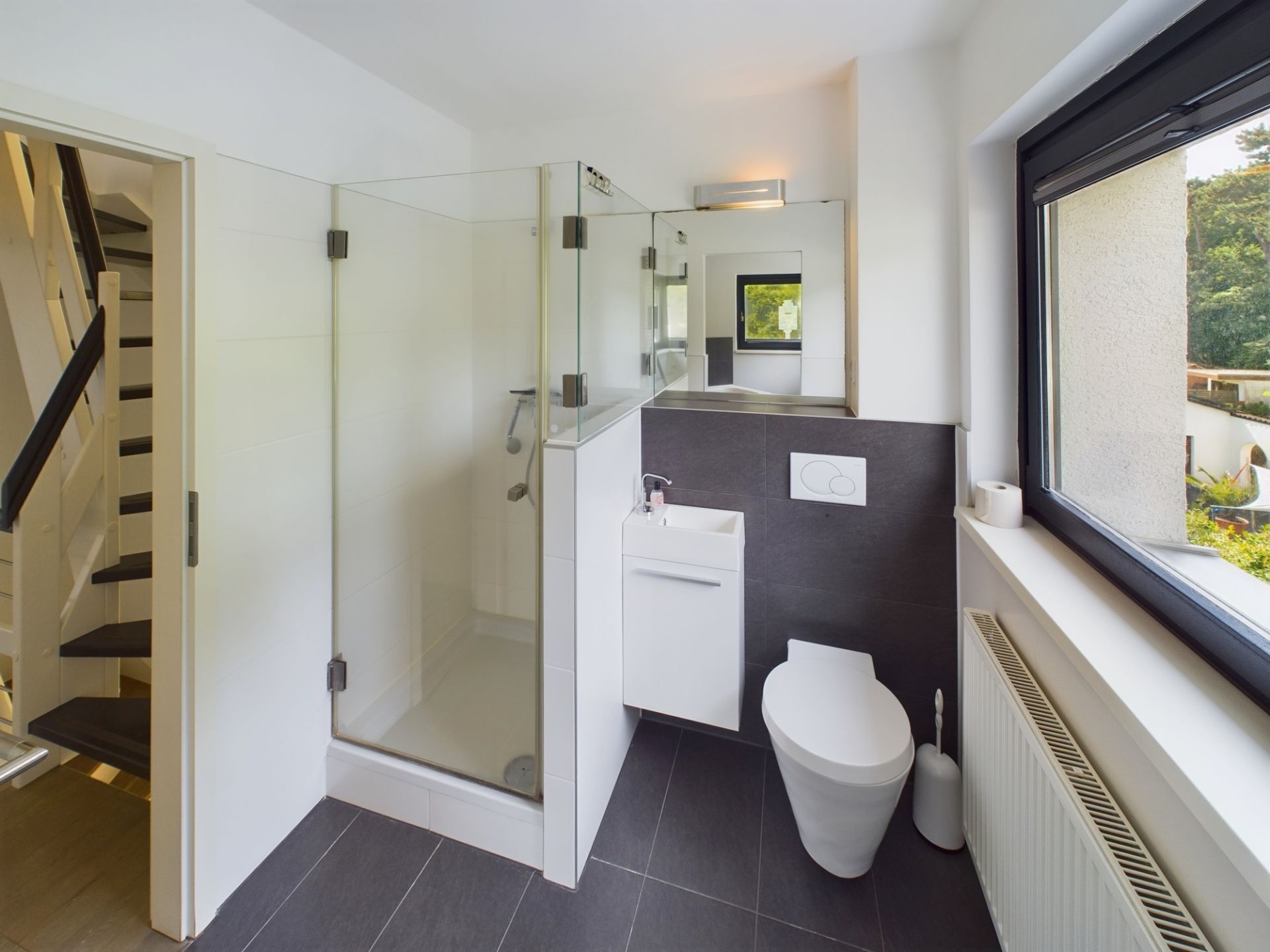 Modernes Badezimmer mit Dusche, WC, Waschbecken und großem Fenster, daneben eine Treppe.