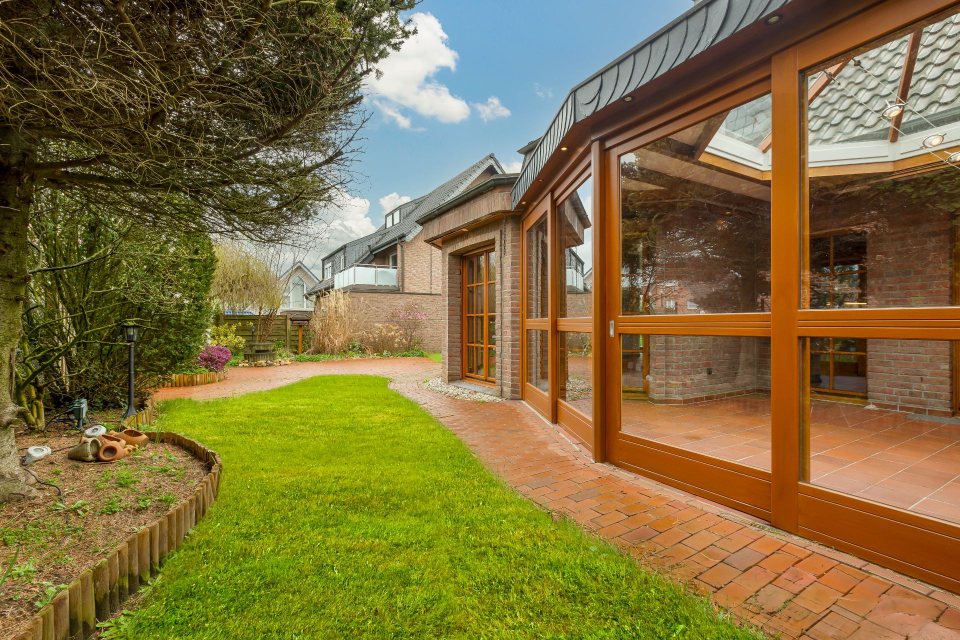 Gepflegter Garten mit Rasen, Weg aus roten Steinen und Wintergarten mit Glasfront.