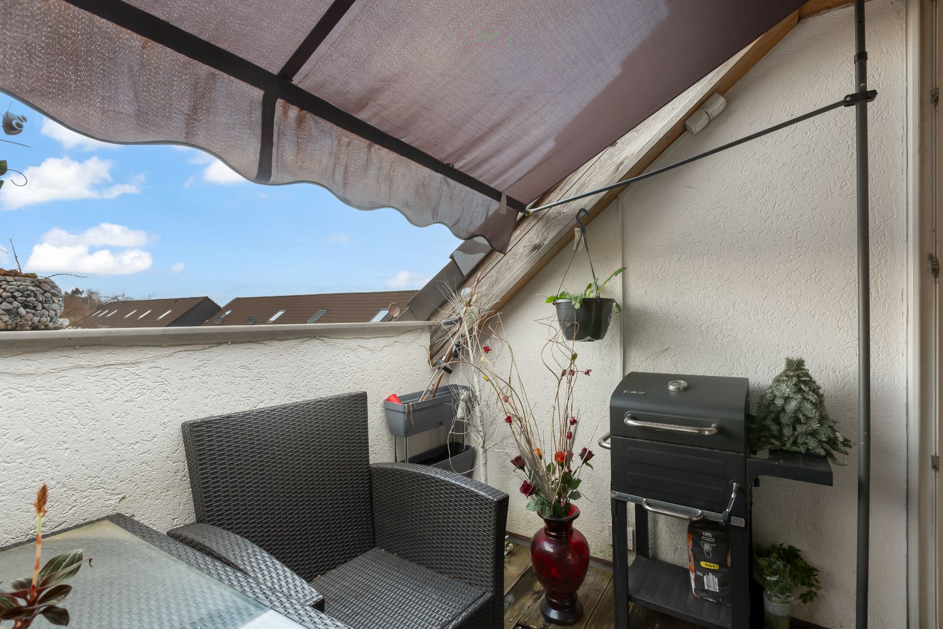Überdachter Balkon mit Sitzmöbeln, Grill, Pflanze im Topf und Blick auf Nachbarhäuser.