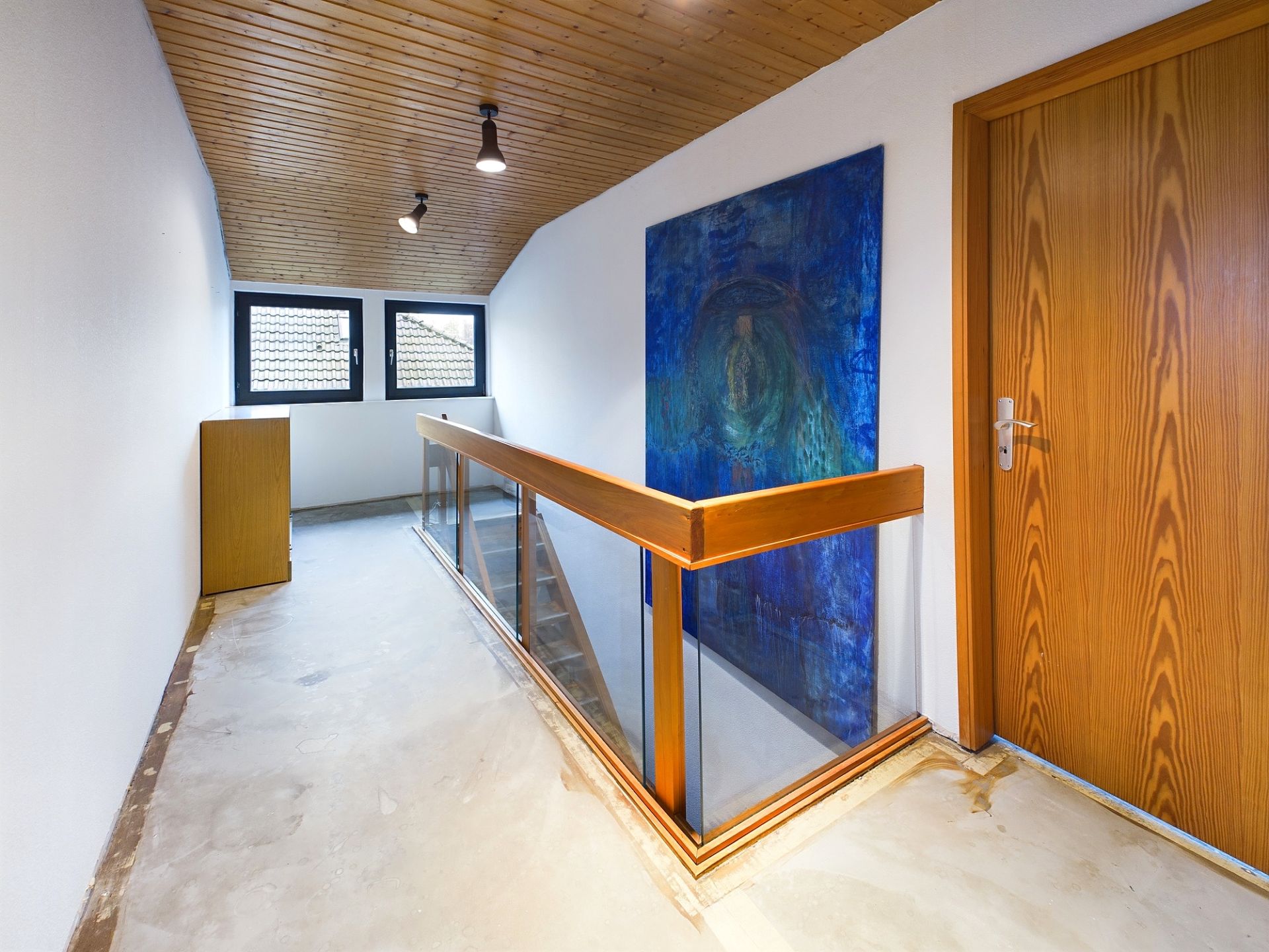 Flur mit Holztür, Glasgeländer, großem blauen Bild und zwei Fenstern am Ende.