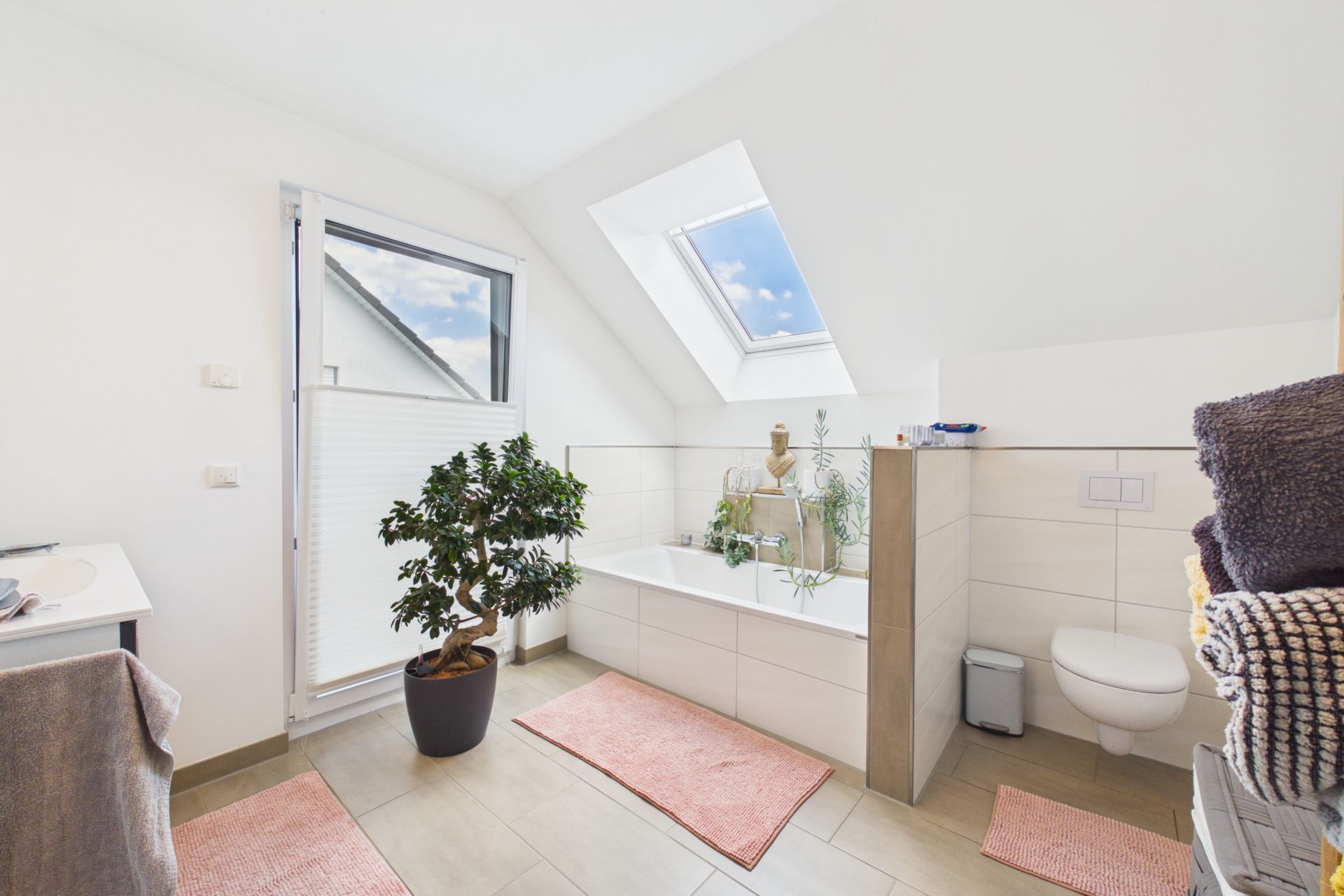 Helles Badezimmer mit Badewanne, WC, Pflanze, zwei Fenstern und rosa Teppichen.