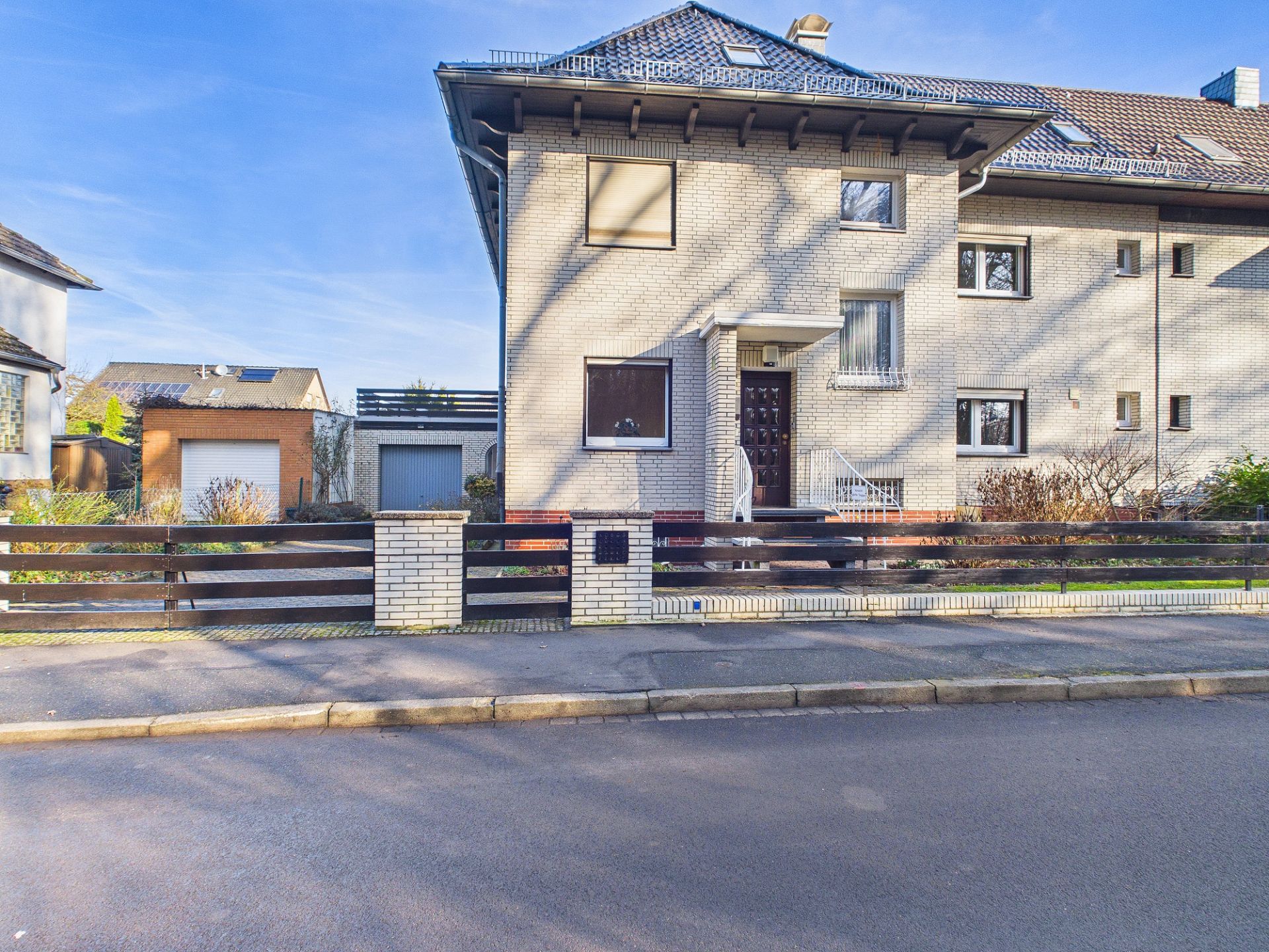 Zweistöckiges Wohnhaus mit hellem Klinker, Vorgarten und Holzzaun an der Straße.