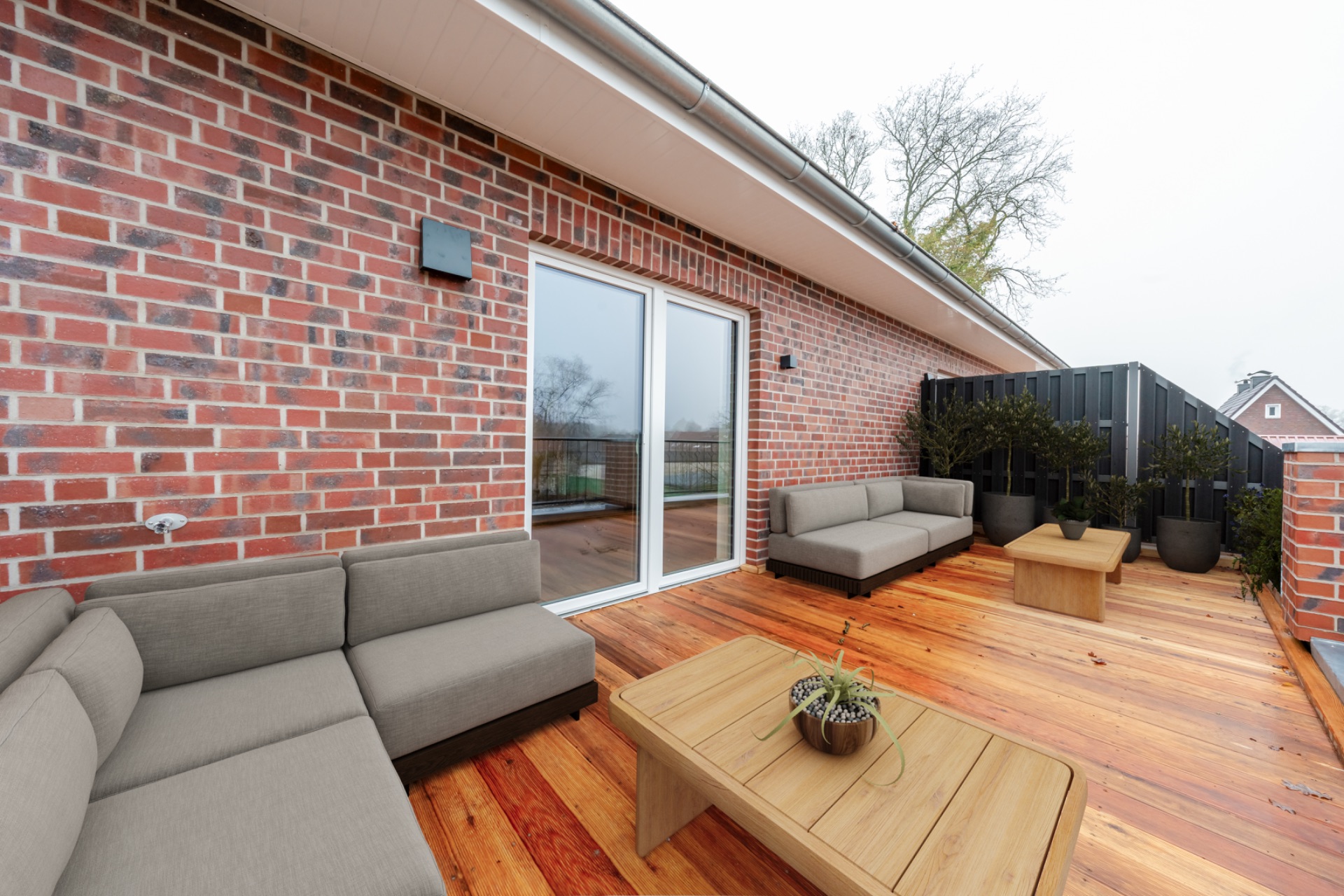 Holzbalkon mit grauen Sofas, Tisch und Pflanzkübeln an einer roten Ziegelwand.