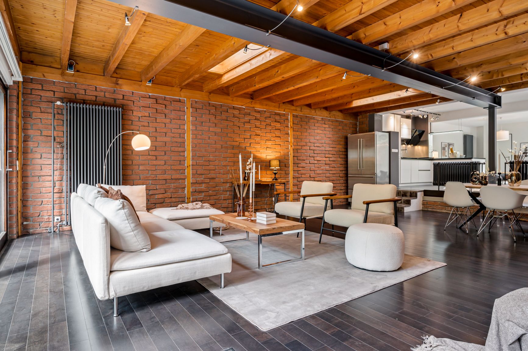 Modernes Wohnzimmer mit Holzbalkendecke, Backsteinwand, Sofa, Sesseln und offener Küche.