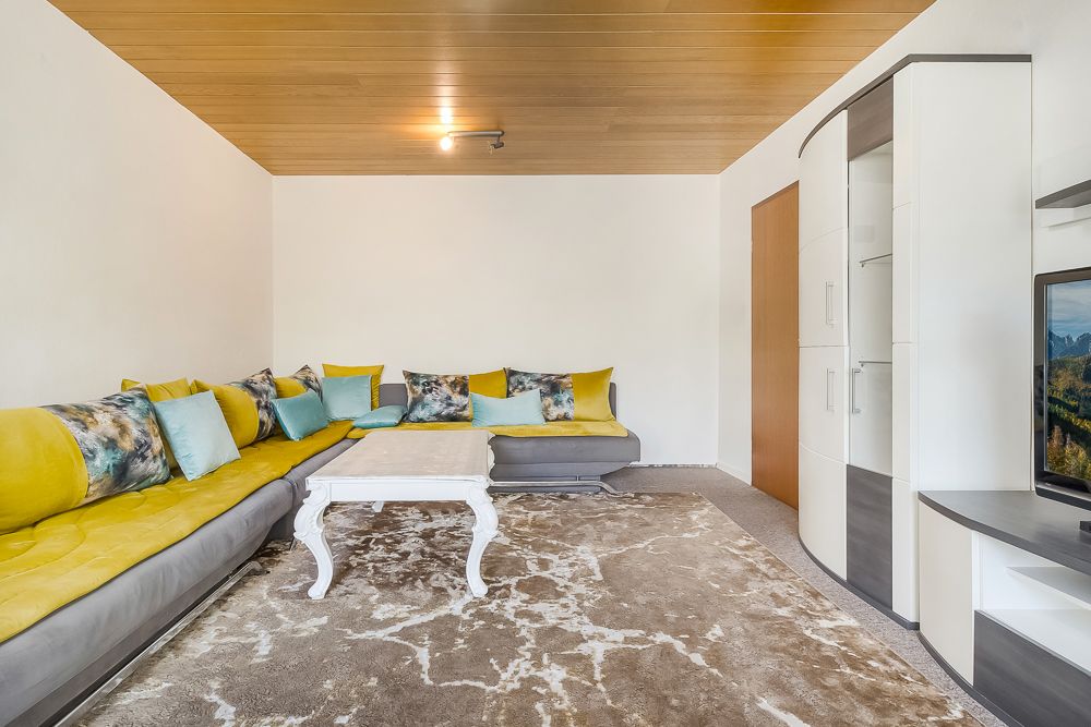 Wohnzimmer mit gelbem Ecksofa, vielen Kissen, Couchtisch und TV-Schrank auf hellem Teppich.