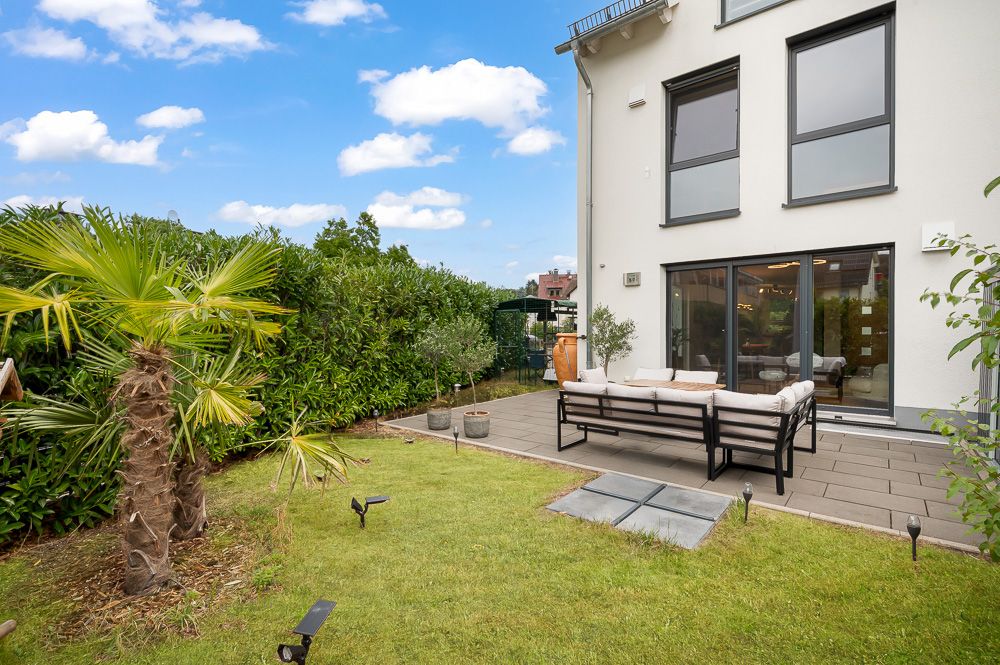 Garten mit Terrasse, Sitzbank, Palmen und großen Fenstern an einem Wohnhaus.