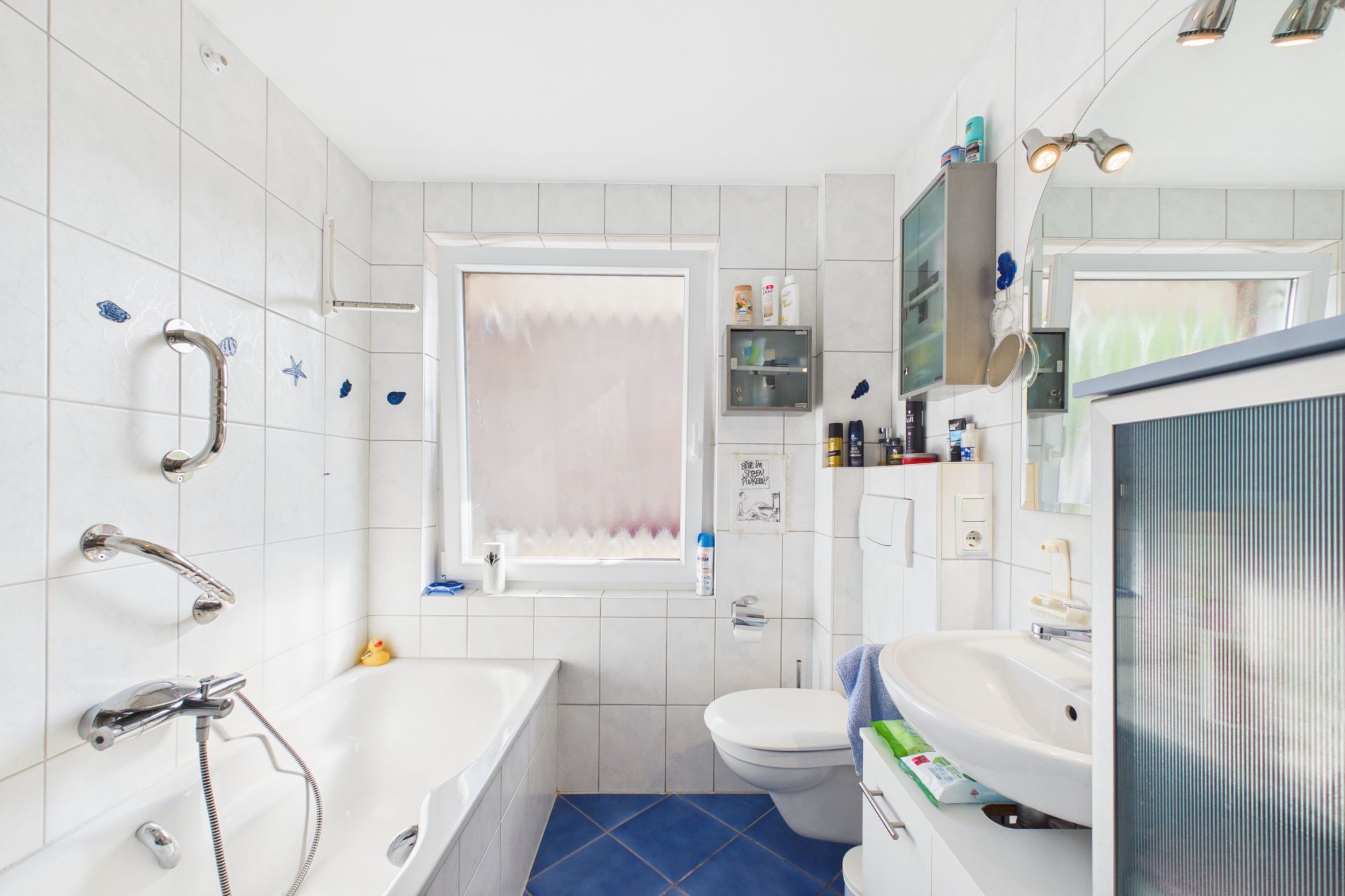 Helles Badezimmer mit Badewanne, Waschbecken, WC und Fenster, alles in weißen Fliesen gehalten.