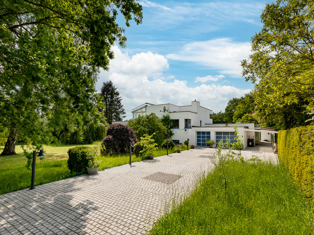 Modernes weißes Wohnhaus mit großer Einfahrt und umliegendem Garten, umgeben von Bäumen.