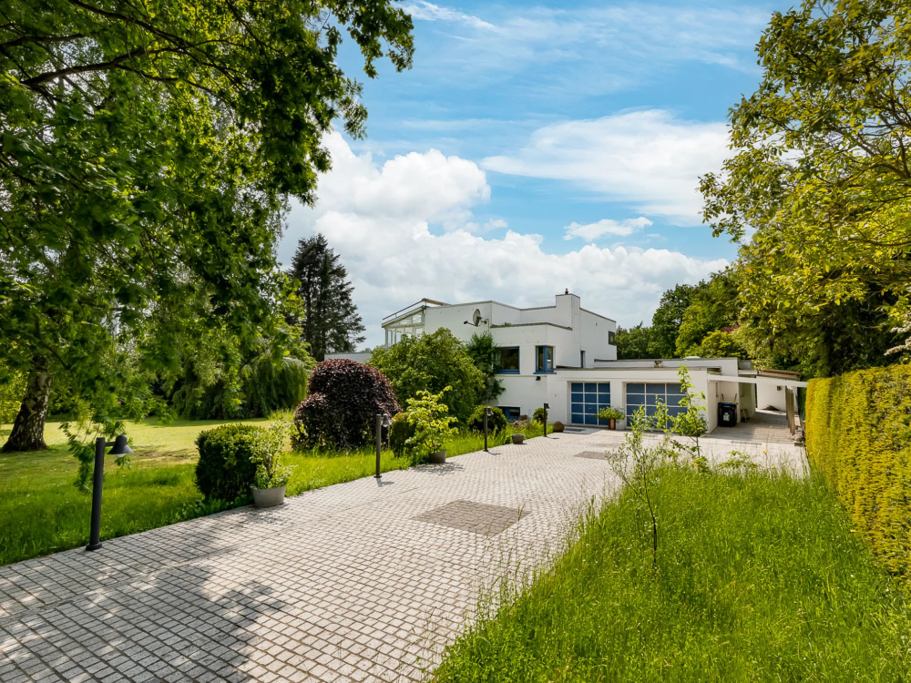 Modernes weißes Wohnhaus mit großer Einfahrt und umliegendem Garten, umgeben von Bäumen.