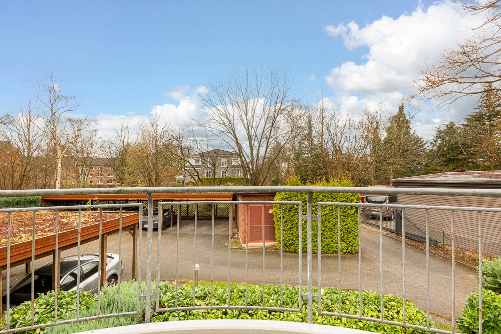 Blick von einem Balkon auf eine Einfahrt mit Carport, Sträuchern und Bäumen im Hintergrund.