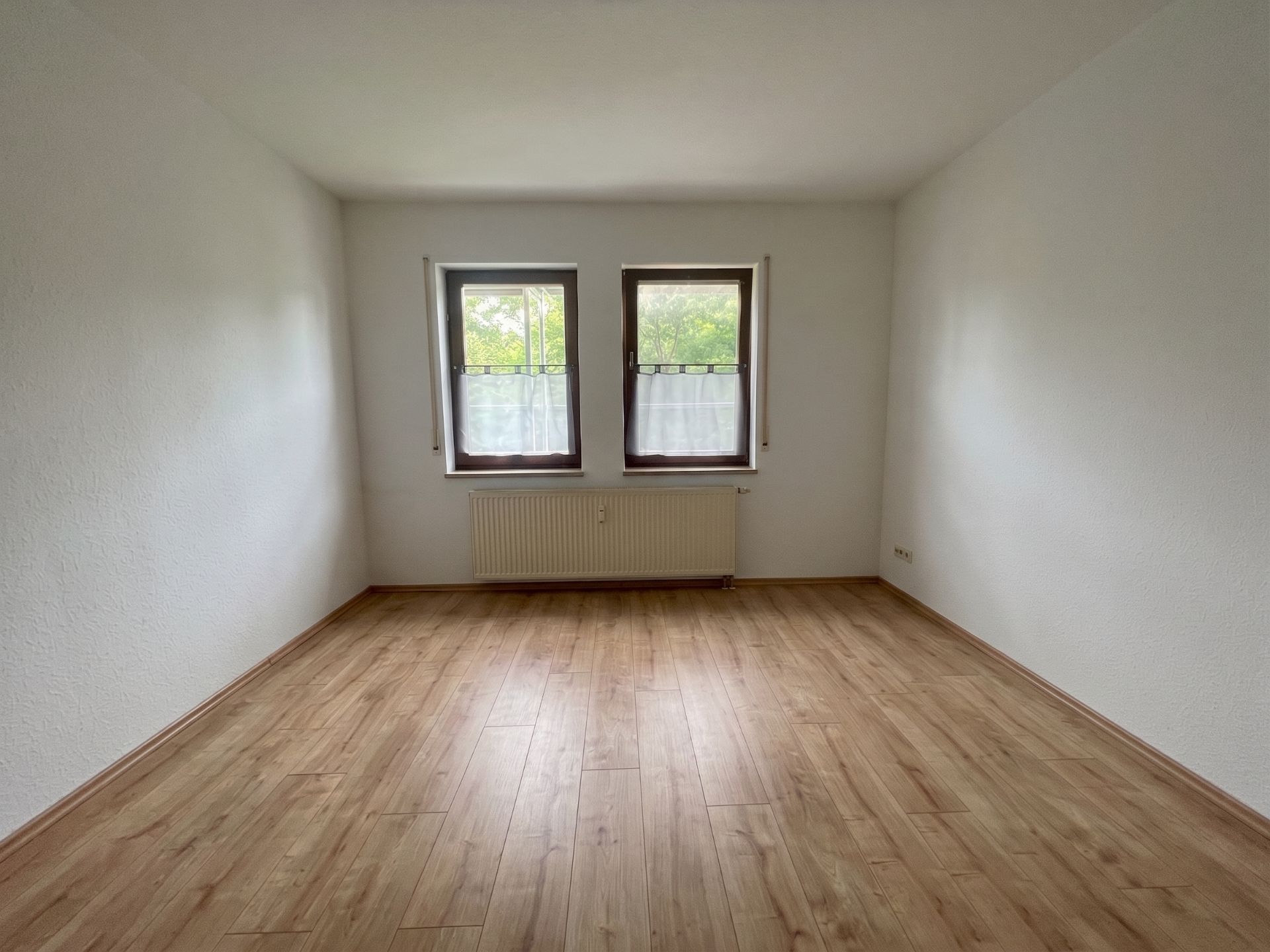 Leerer Raum mit hellem Holzfußboden und zwei Fenstern mit weißen Vorhängen.